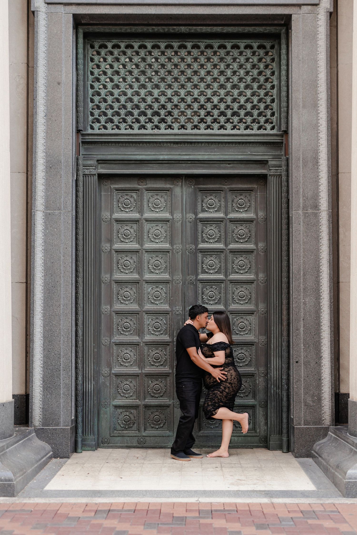 A couple embraces in front of an ornate gray door with geometric patterns.