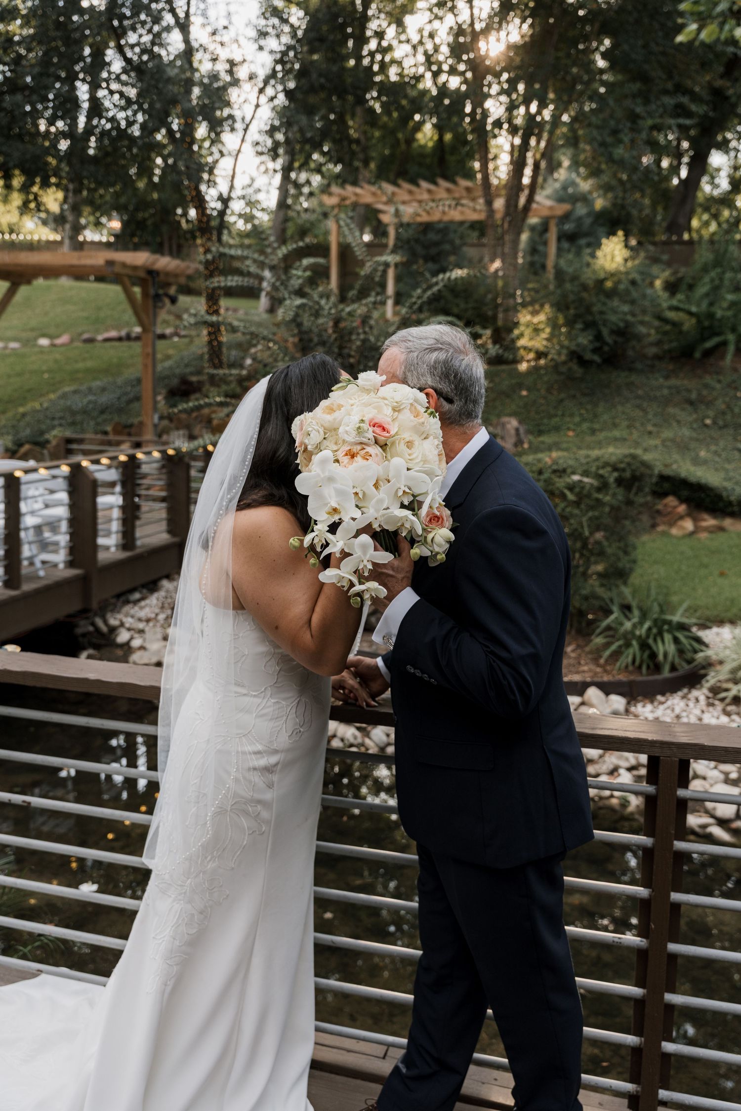 Bride in white gown and veil shares a tender moment with older man on wooden bridge, bouquet of white flowers visible.