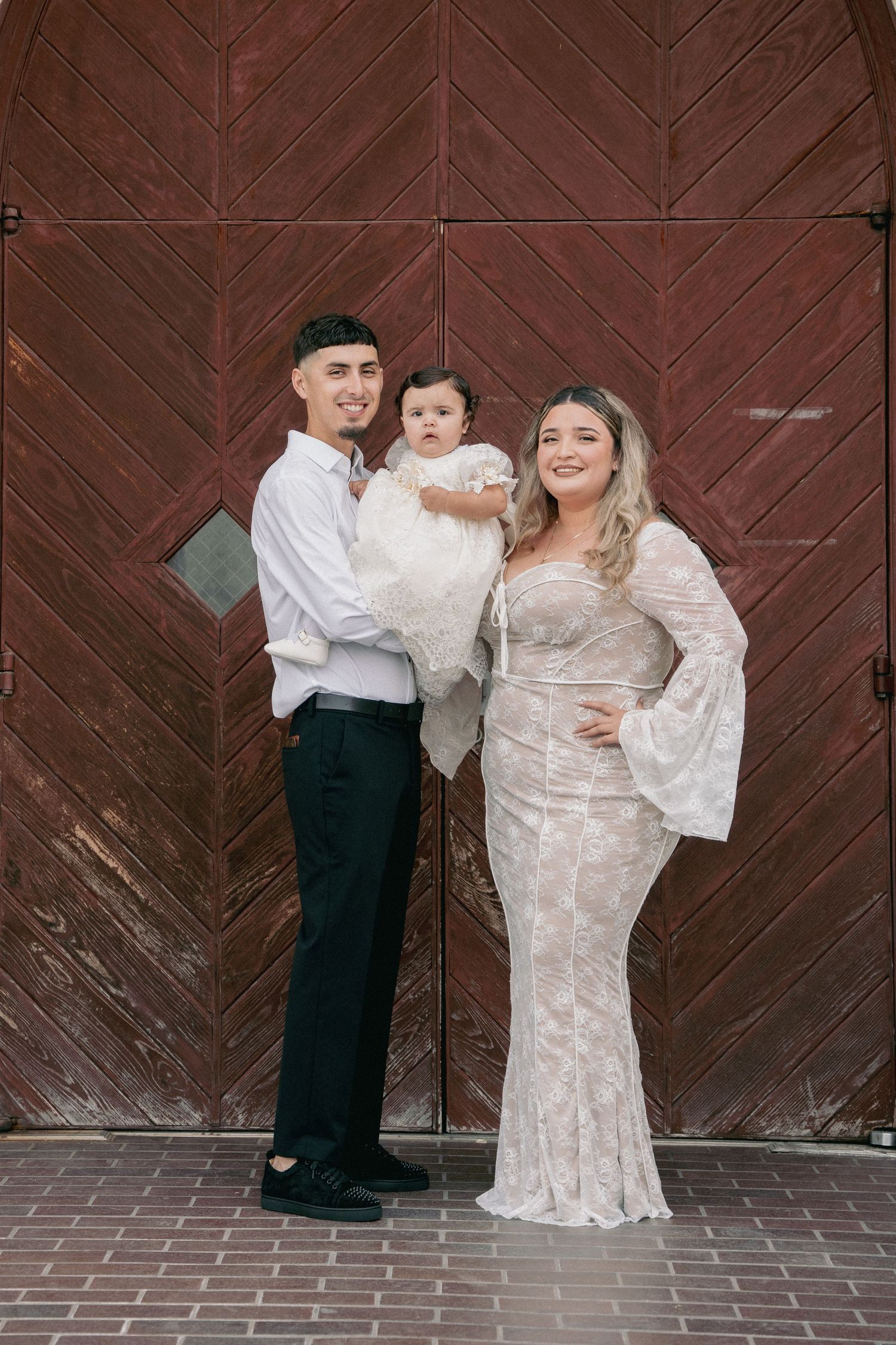 A couple in elegant white attire poses with a child in front of a wooden church door.