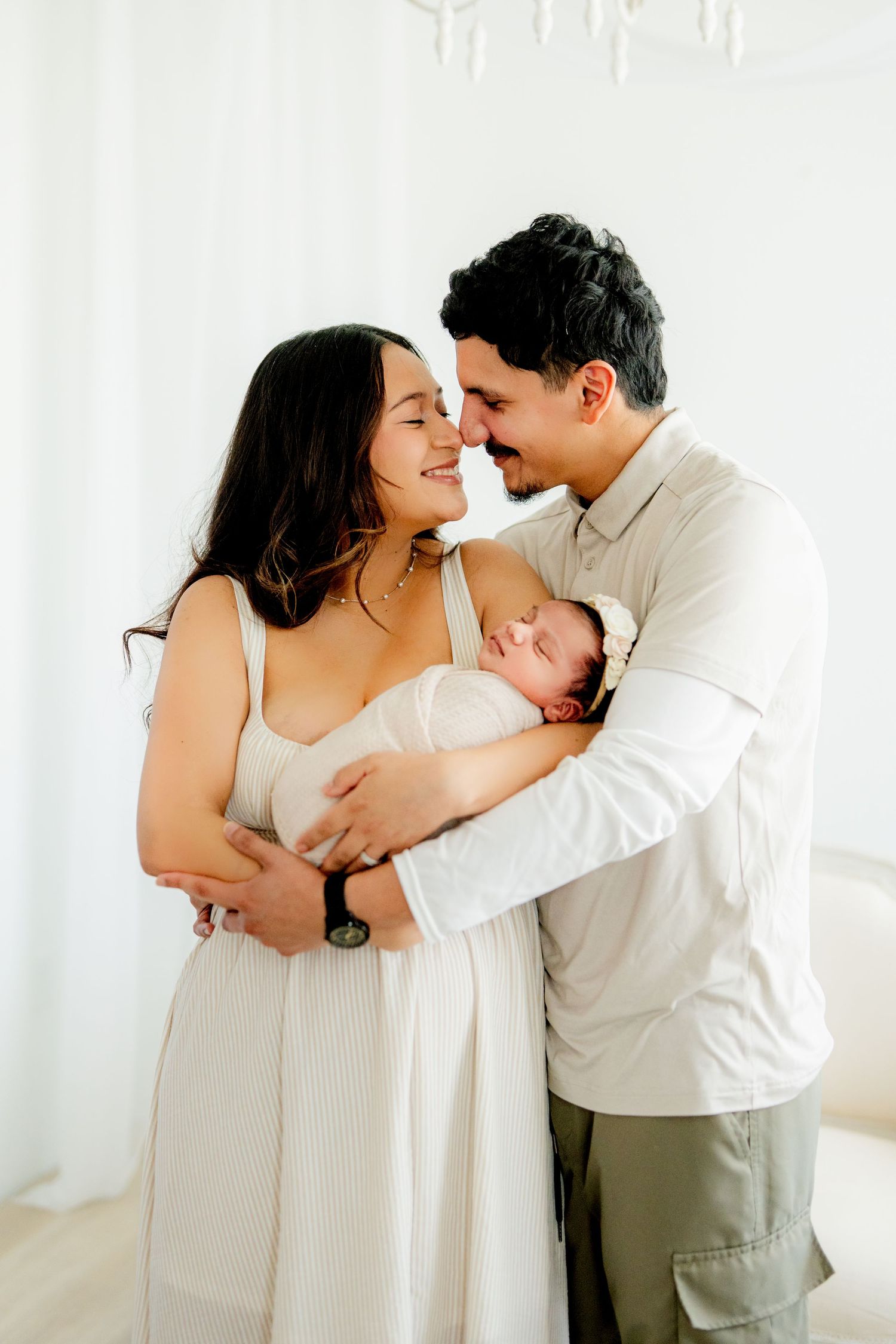 A loving couple shares an intimate moment while holding their sleeping newborn baby in a bright, sunlit room.
