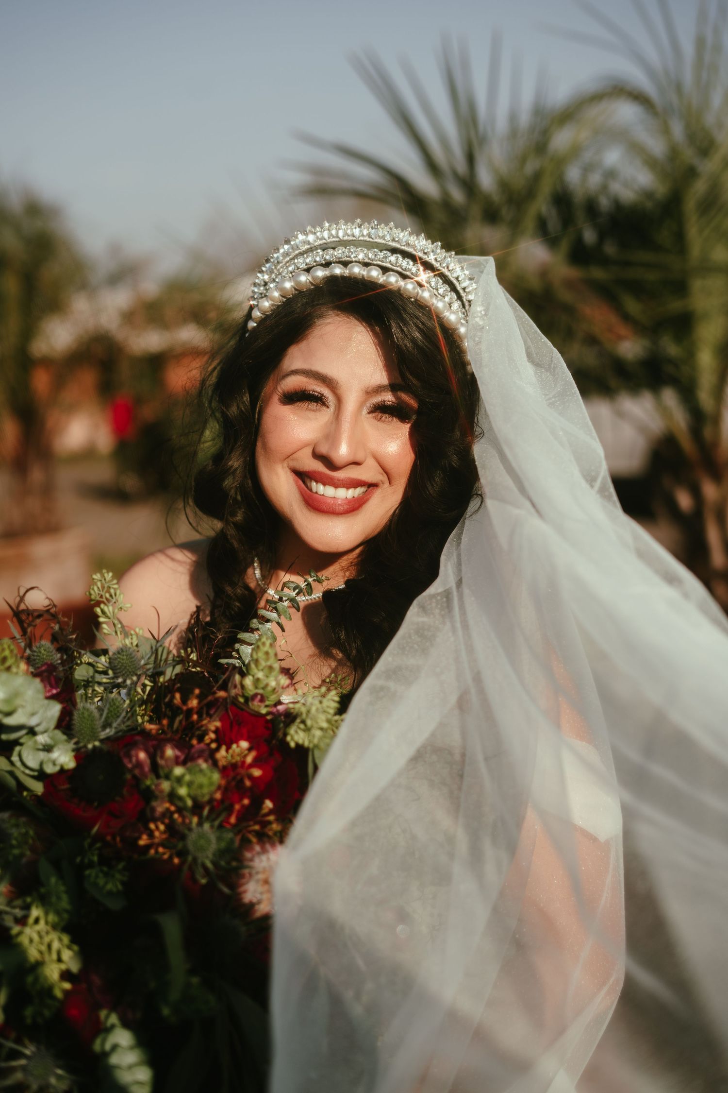 A radiant bride wearing a crystal crown and veil smiles while holding a colorful bouquet.