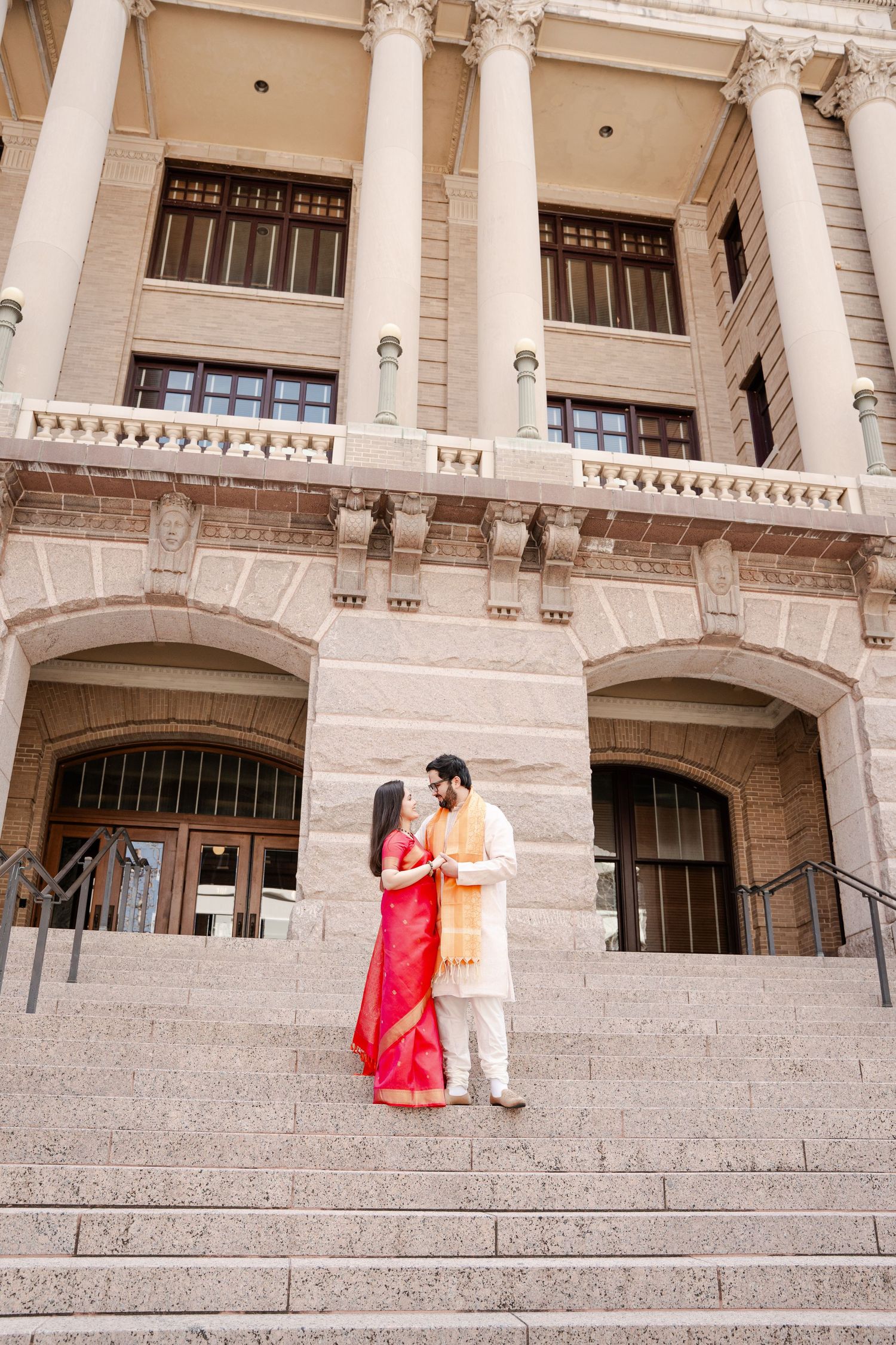 A couple in traditional Indian wedding attire stands on grand stone steps of a classical building.