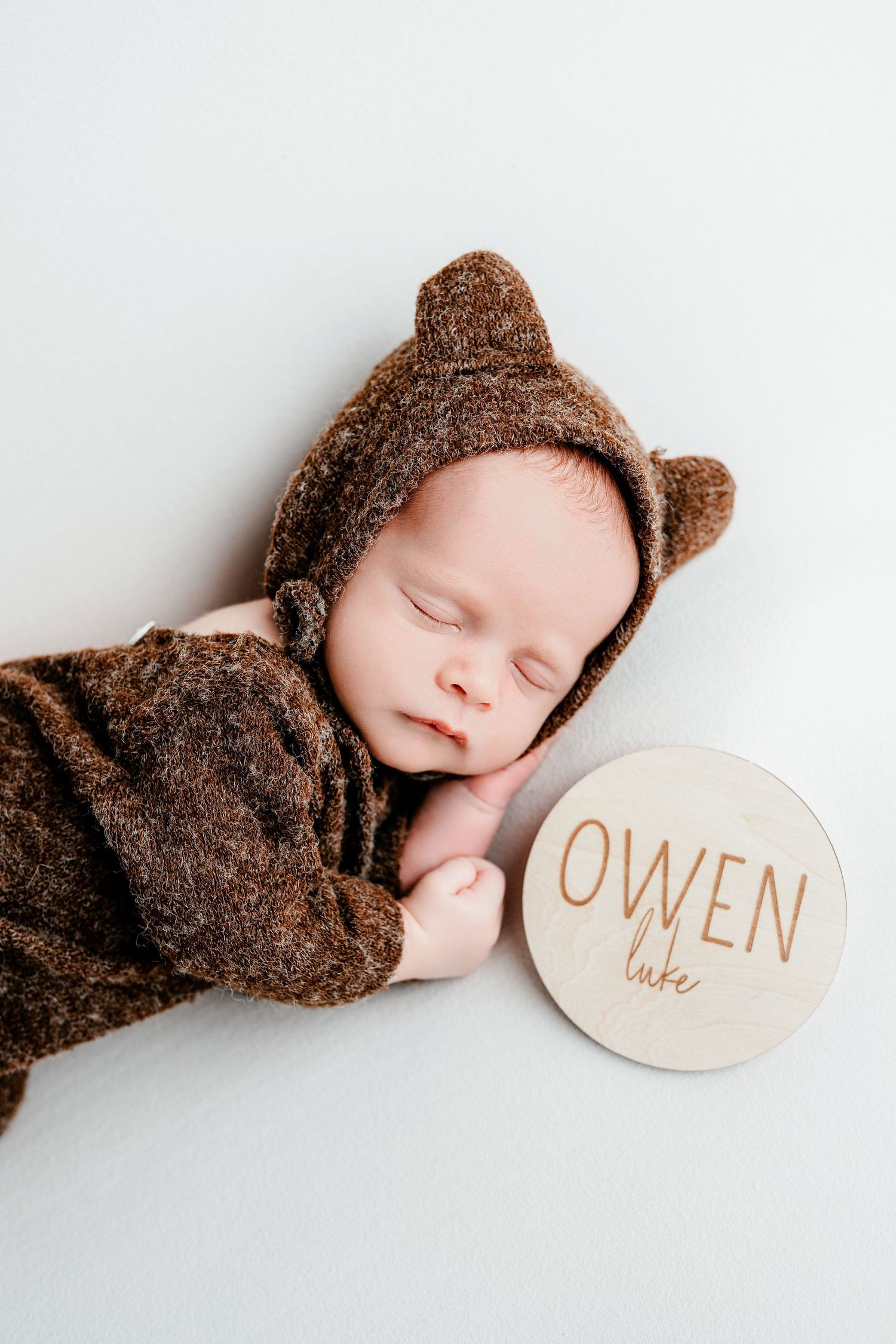 Sleeping newborn in cozy brown bear outfit lying next to wooden name sign reading Owen.
