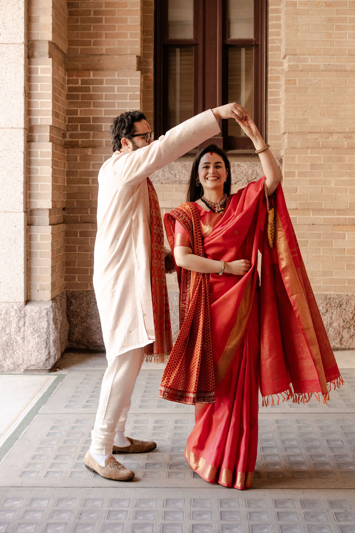 A couple in traditional Indian attire dancing together in a brick archway, with the woman wearing a vibrant red saree.
