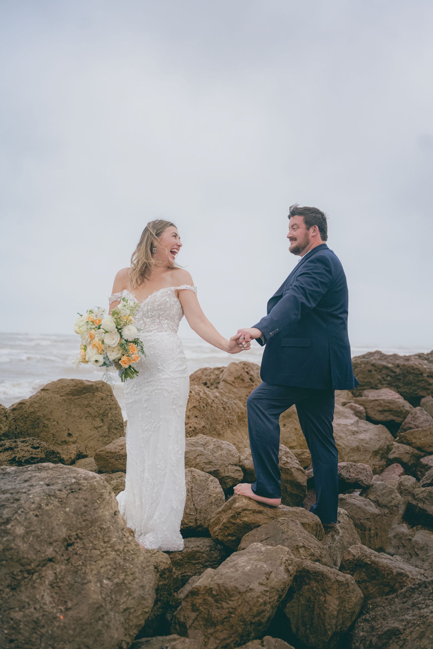 A couple shares a joyful moment together on rocky coastal terrain during their wedding photo session.