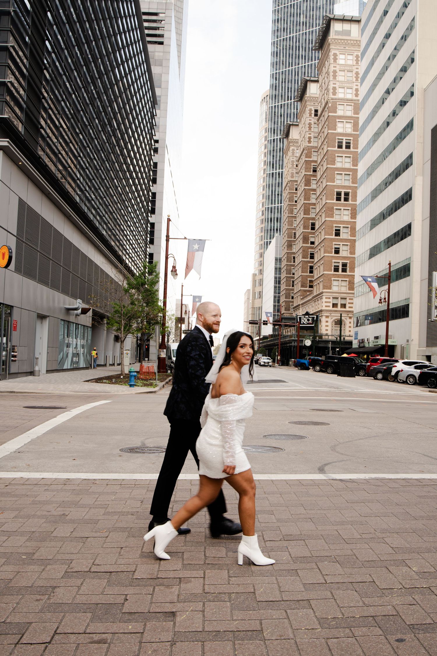 A newlywed couple dances playfully on a city street with modern buildings in the background.