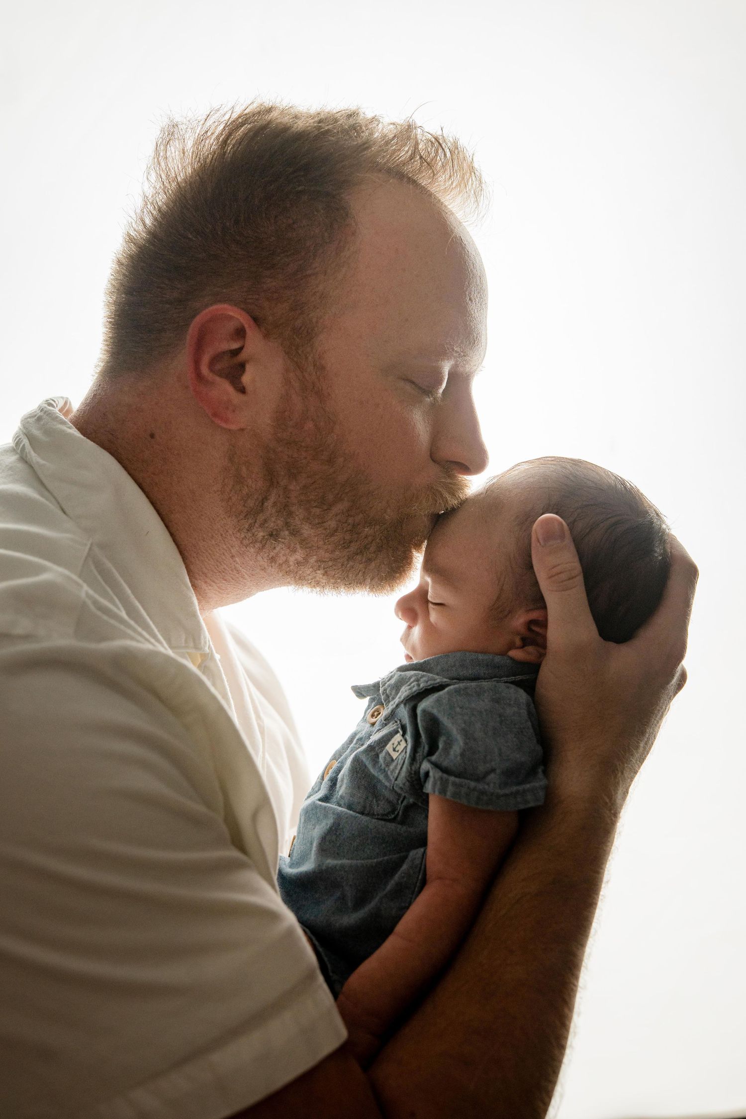 A tender moment between father and newborn captured in soft backlit glow as the dad gently kisses his baby's head.