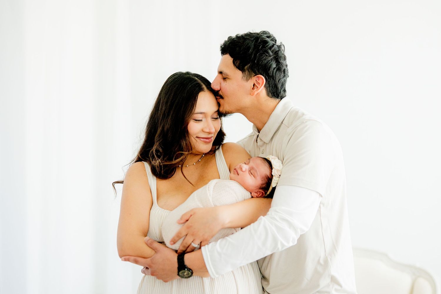 A tender family moment as parents cradle their newborn baby against a bright white background.