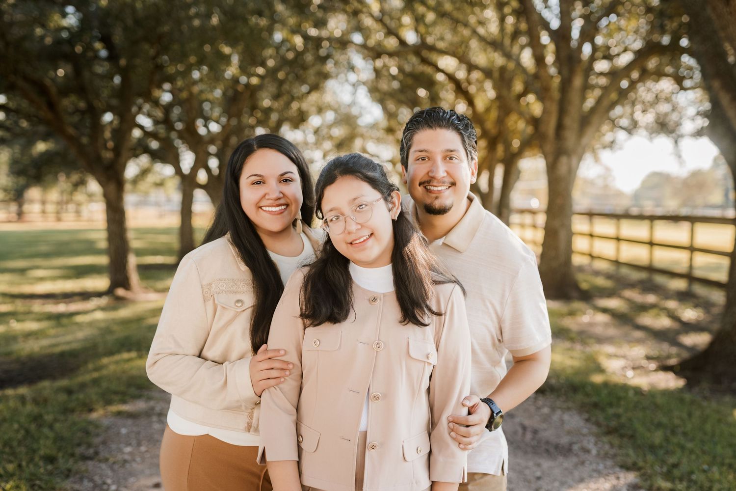 A family group in coordinated white and beige outfits poses outdoors under sunlit trees.
