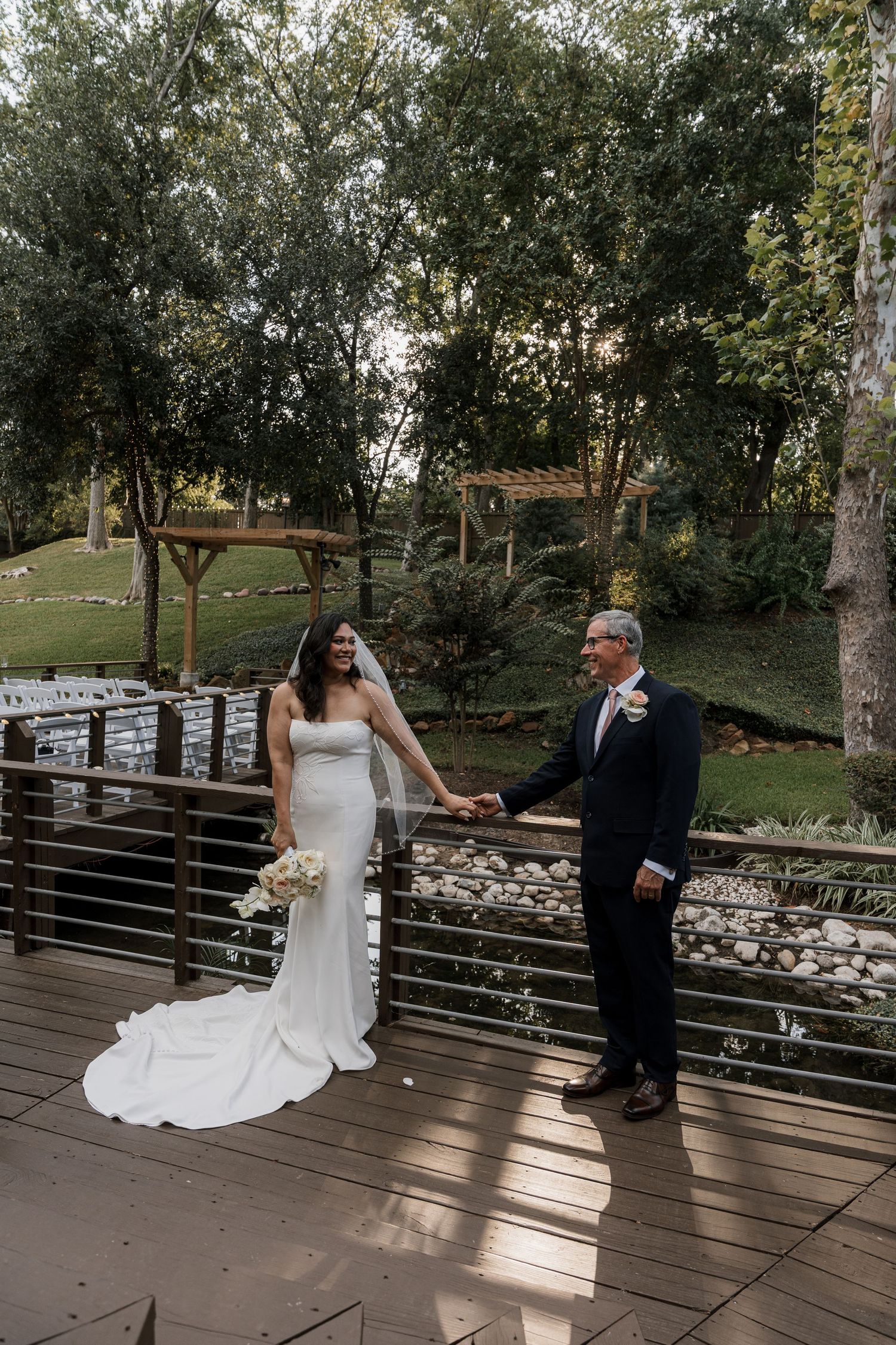 Bride in white gown holds hands with groom in navy suit on wooden deck surrounded by lush greenery at outdoor wedding venue.