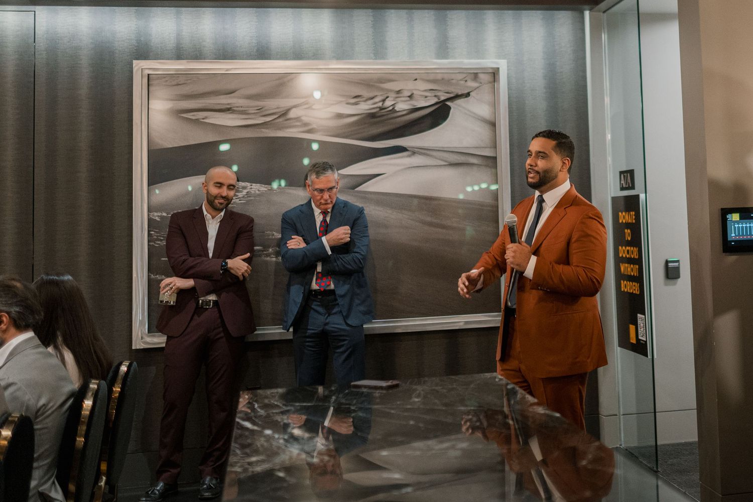 Three well-dressed men in suits socializing in a modern lobby with abstract artwork on the wall.