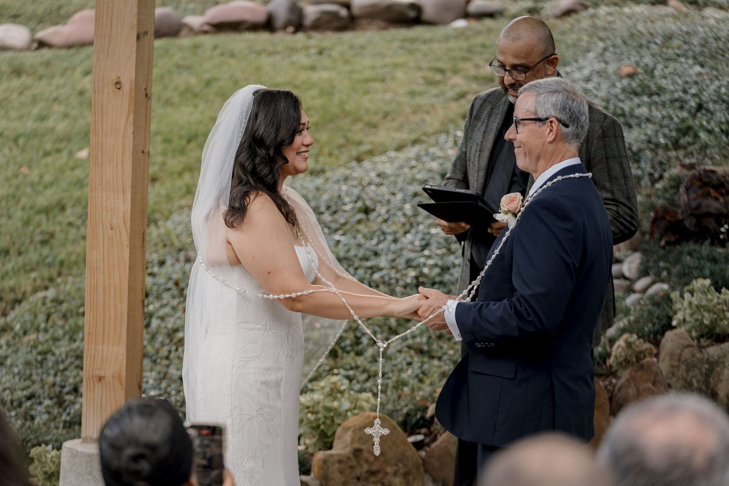 Bride and groom exchange vows outdoors at a rustic wedding ceremony with an officiant holding a book.