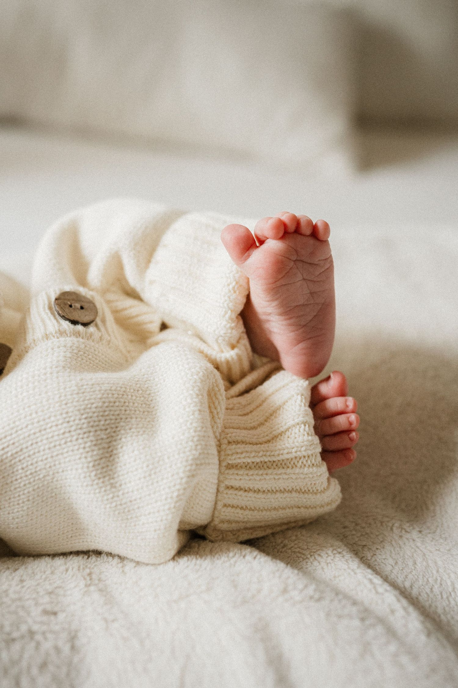 Tiny baby feet peeking out from cozy white knit sweater and blanket in soft natural lighting.