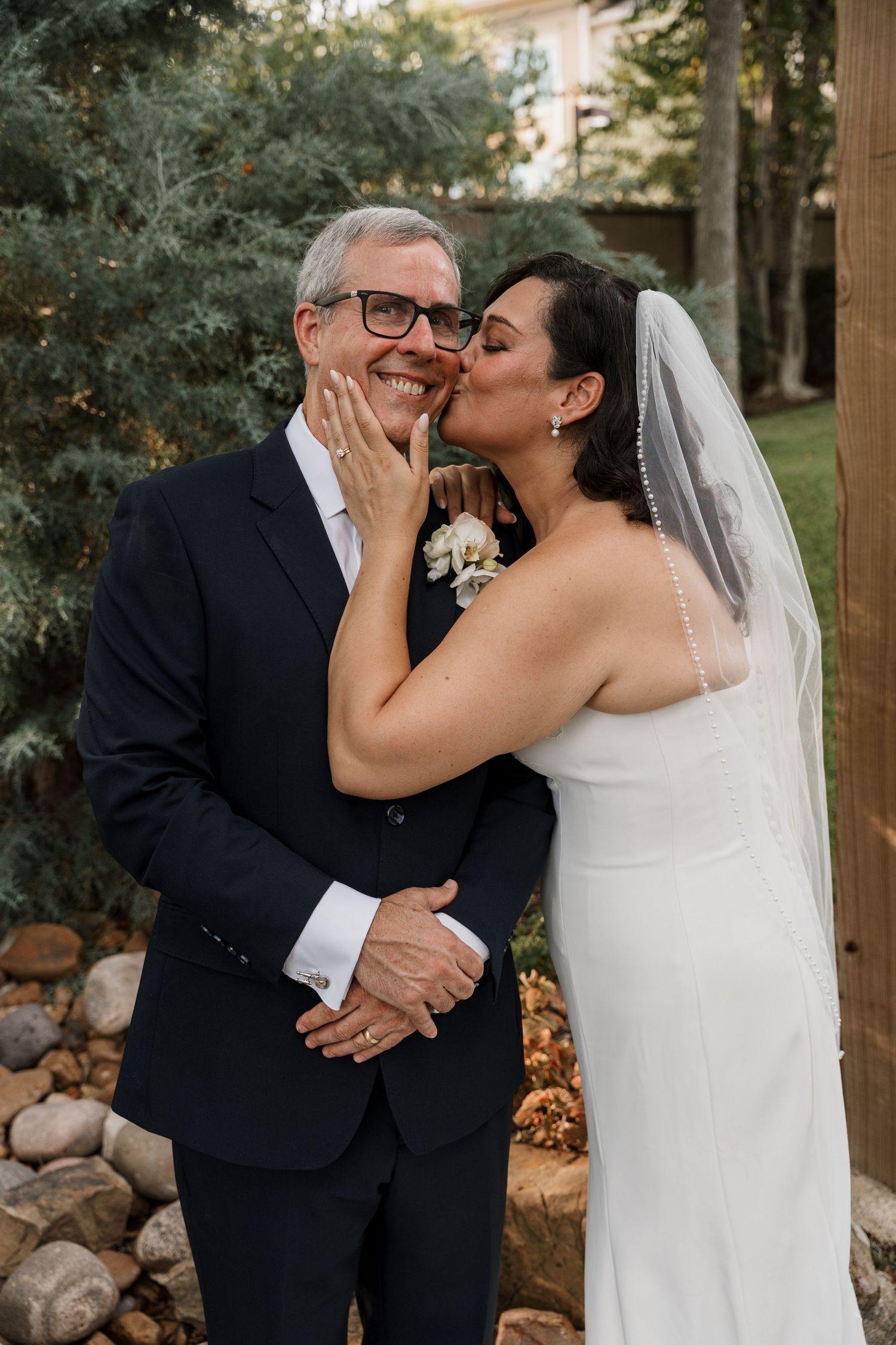 Bride in white gown and veil kisses groom in navy suit on cheek outdoors at wedding ceremony.