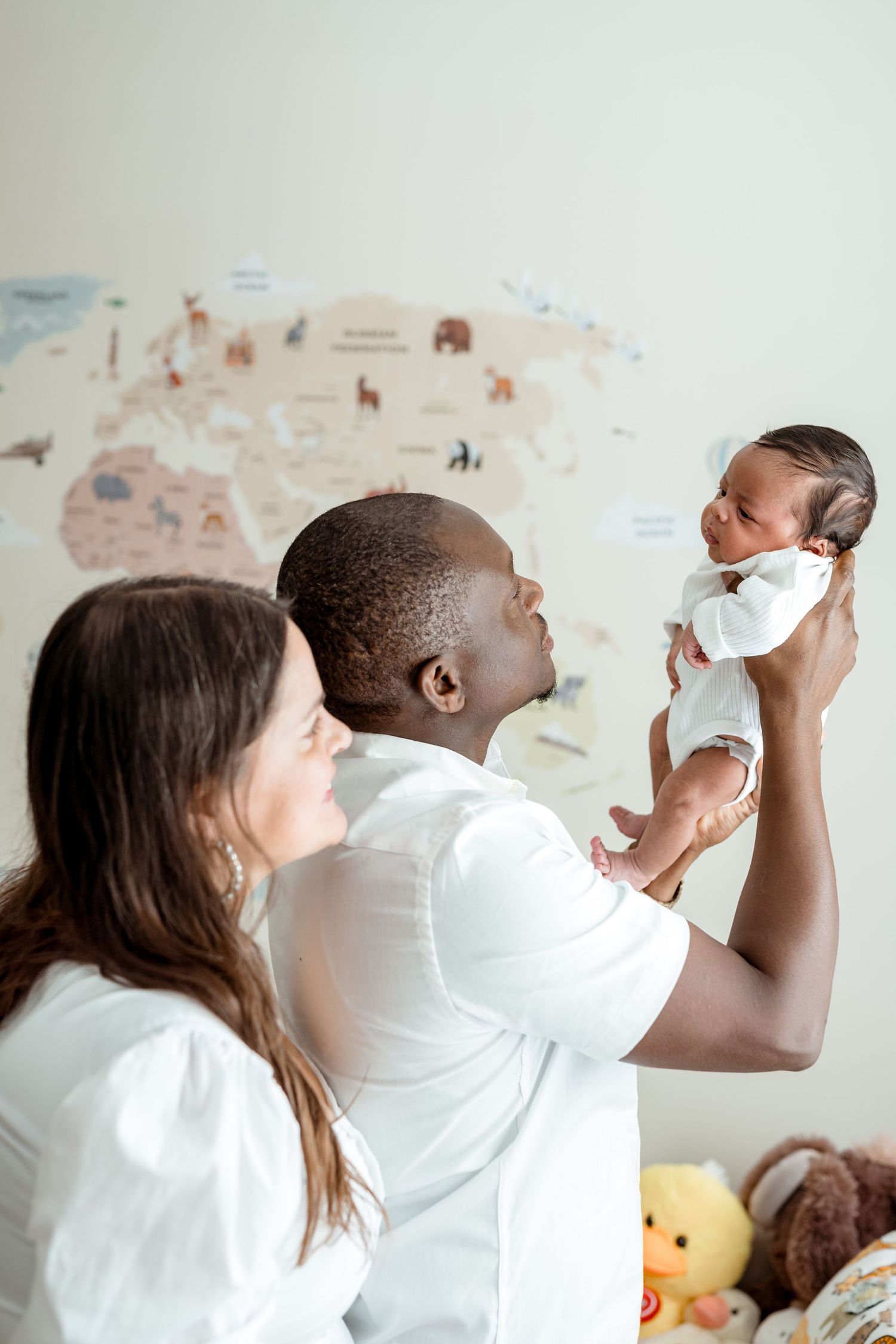 Parents in white outfits holding newborn baby in nursery with world map wall decor.
