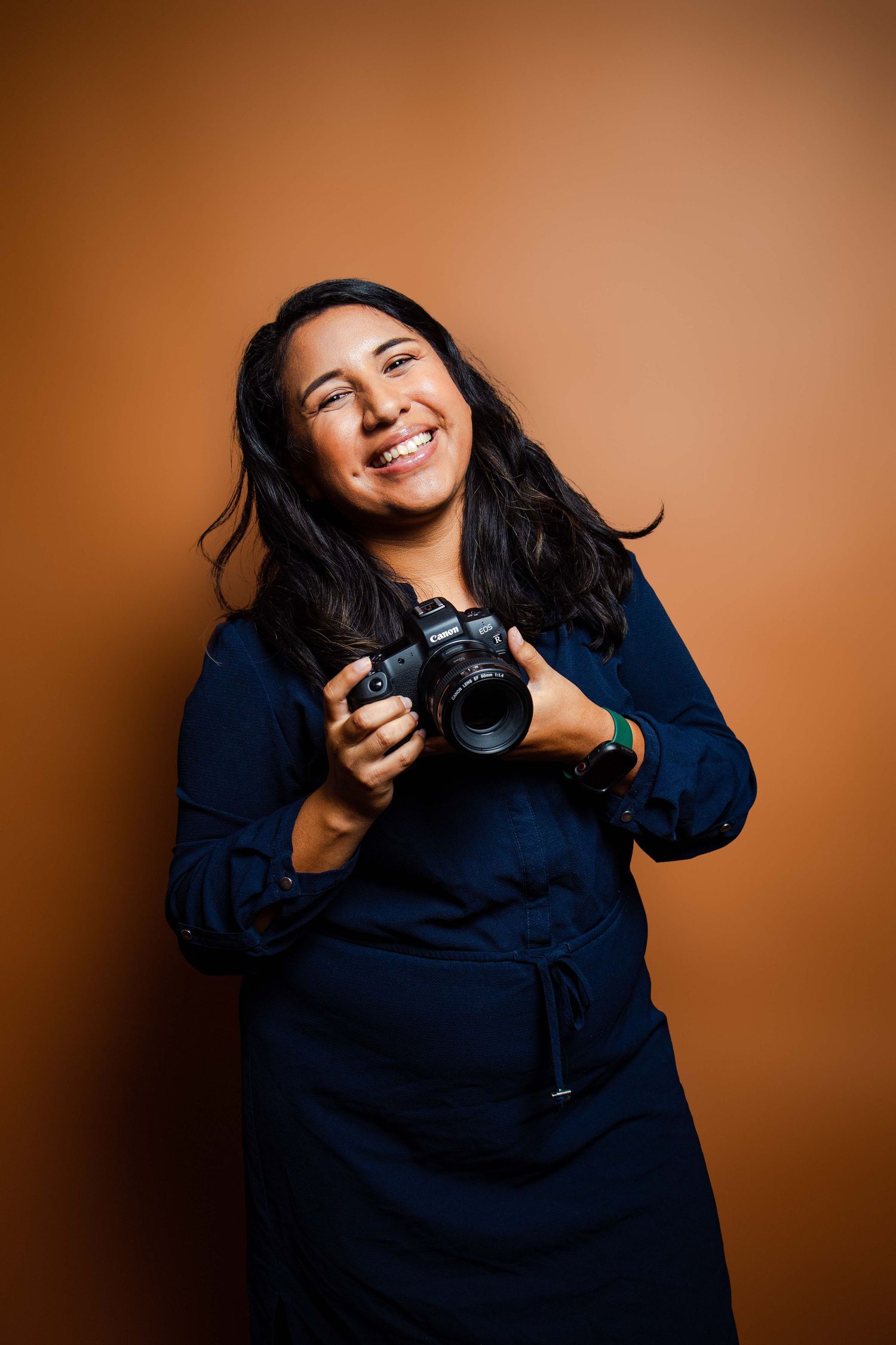A photographer in a navy dress holding a professional camera against an orange background.
