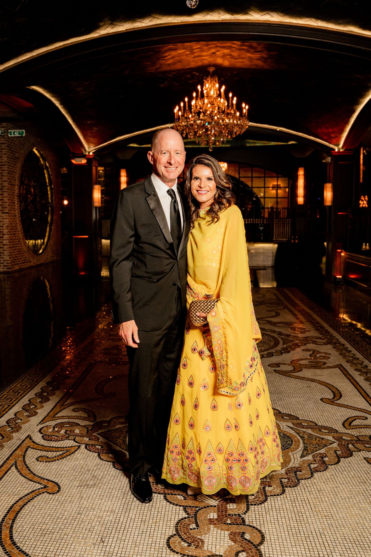 Couple posing in elegant venue, man in dark suit, woman in yellow embroidered lehenga, chandelier backdrop.