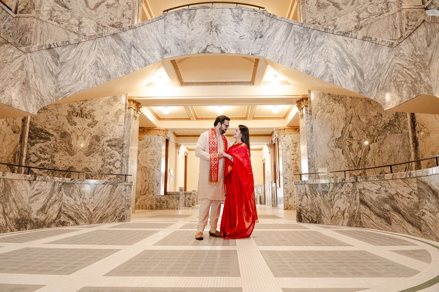 A couple in traditional Indian wedding attire poses under a marble archway in an elegant building.