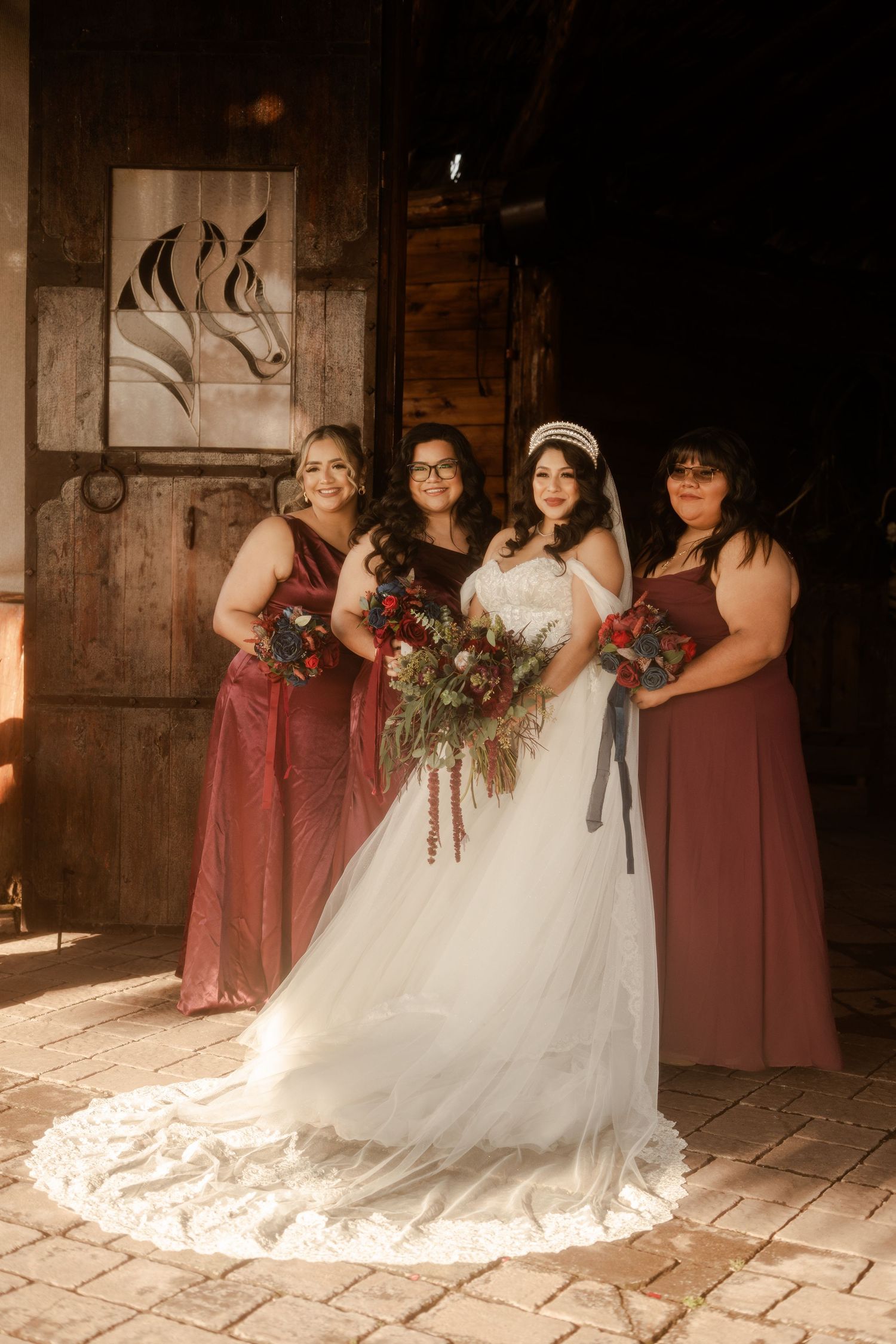 A bridal party poses in a rustic barn setting with bridesmaids in burgundy dresses and white wedding gown.