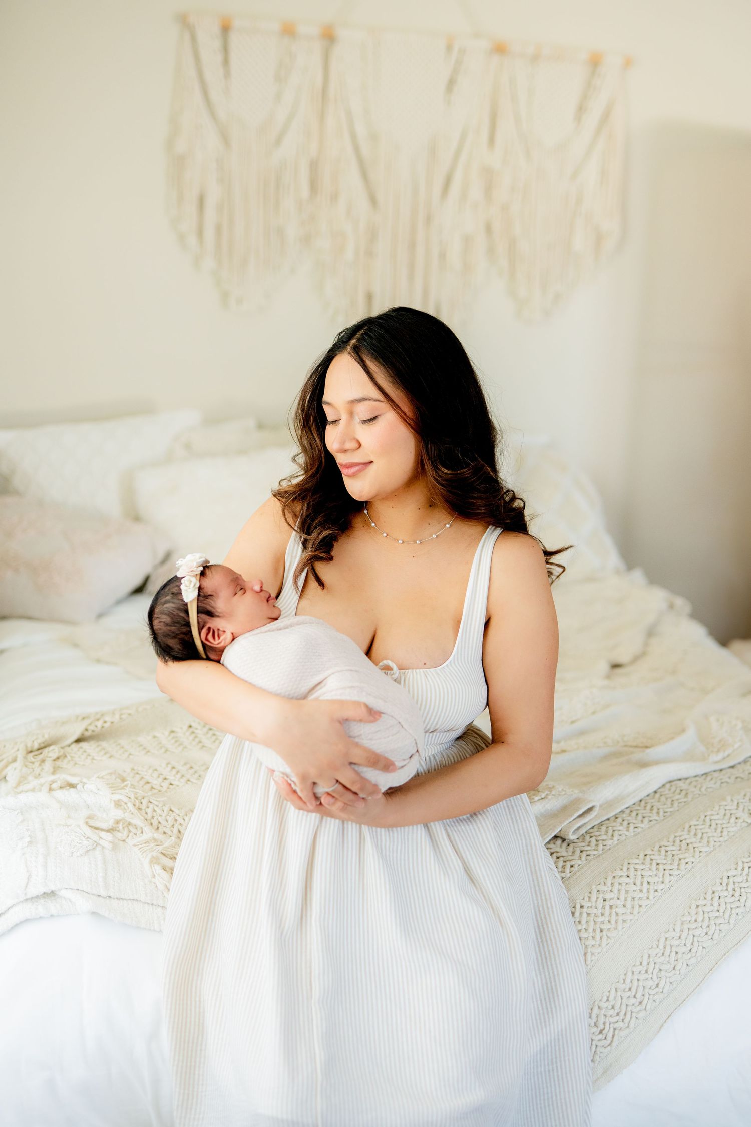 A tender moment between mother and newborn baby on a white bed with macrame wall decor in the background.