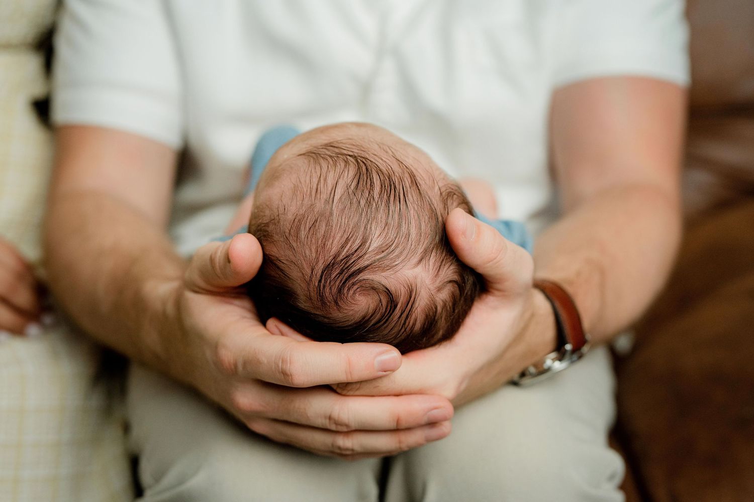 Adult hands gently cradle the back of a newborn baby's head in an intimate close-up photograph.