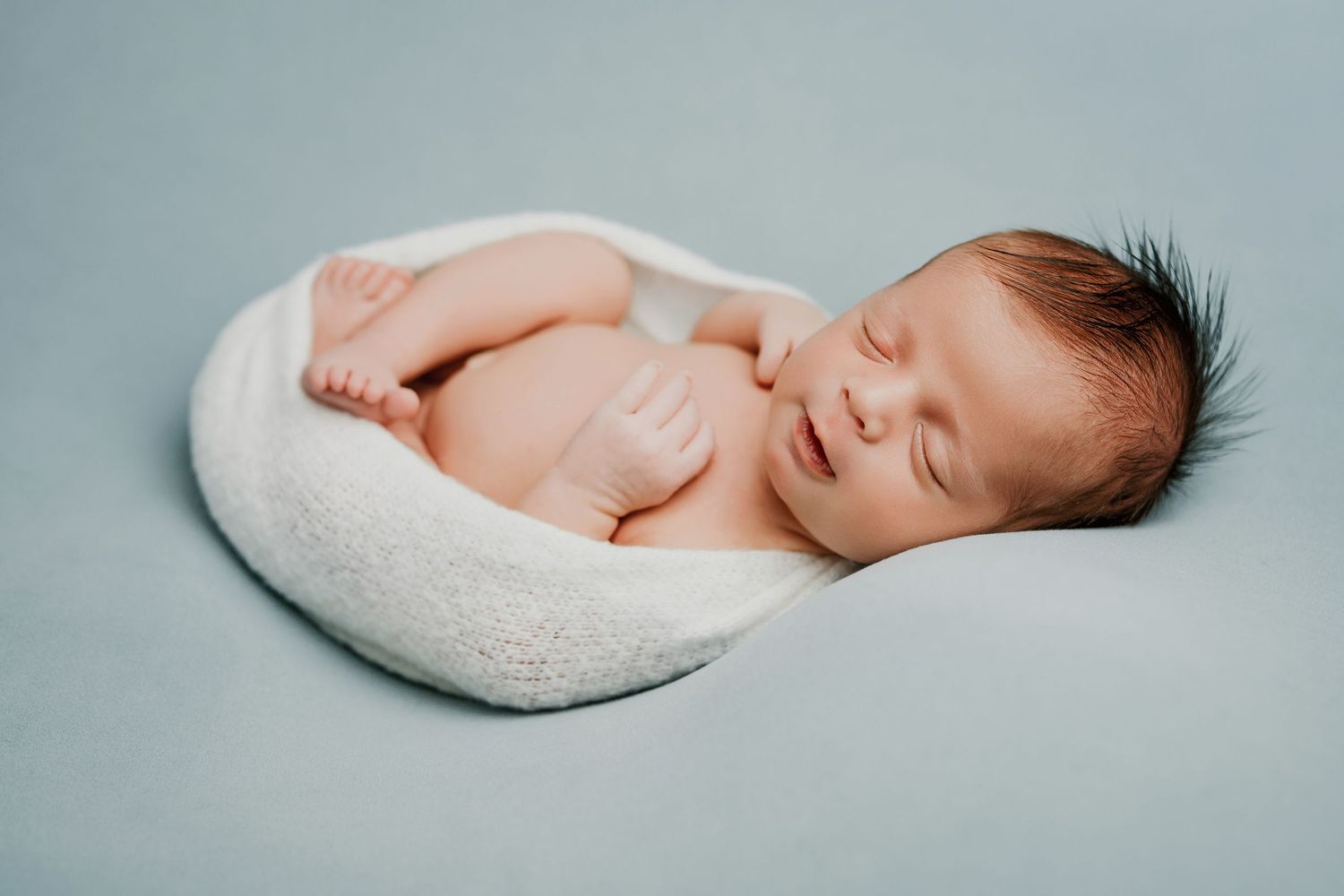 A sleeping newborn baby swaddled in white fabric rests peacefully against a light blue background.