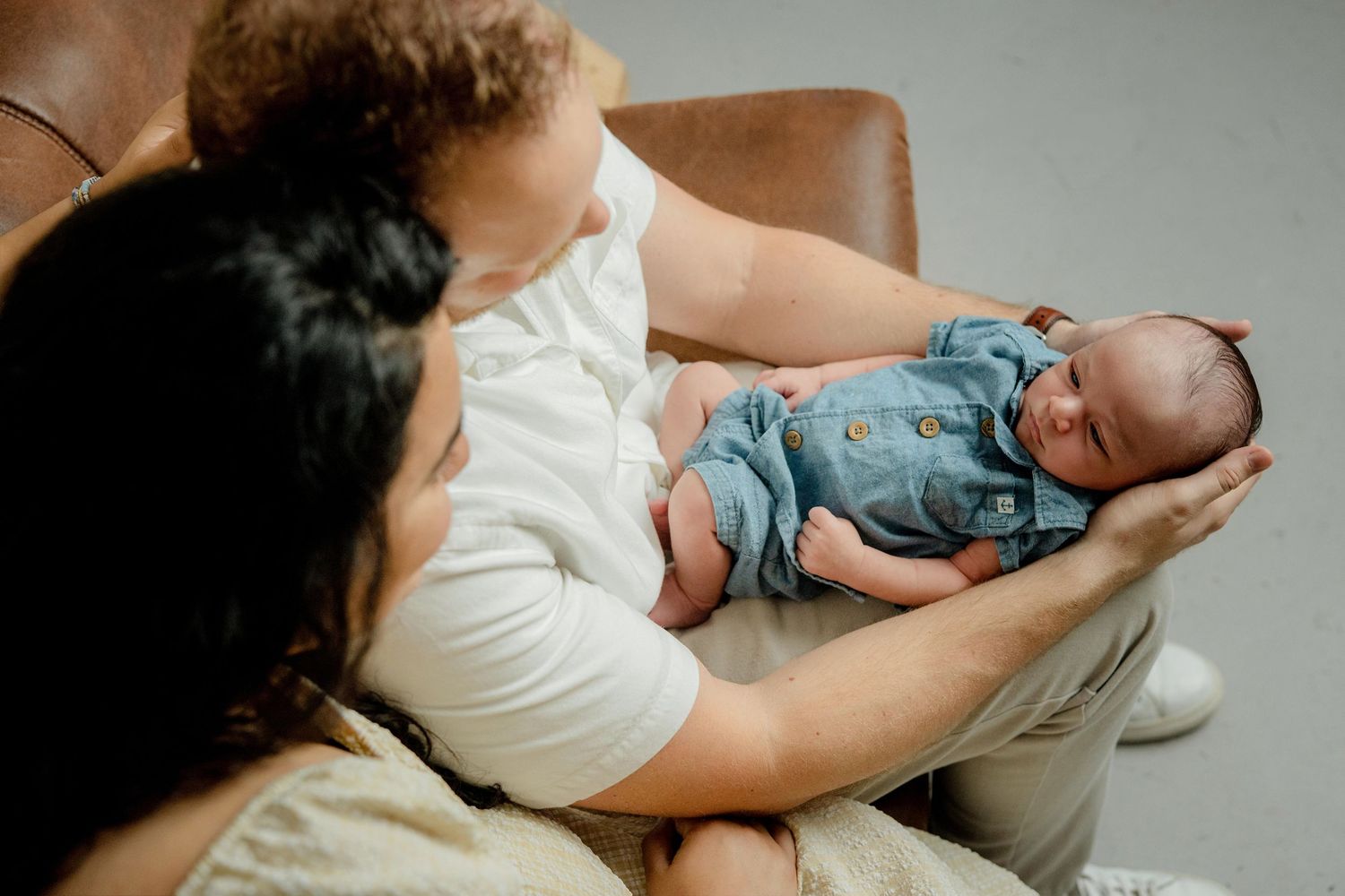 Parent cradling a sleeping infant wearing denim overalls while sitting in a brown leather chair.