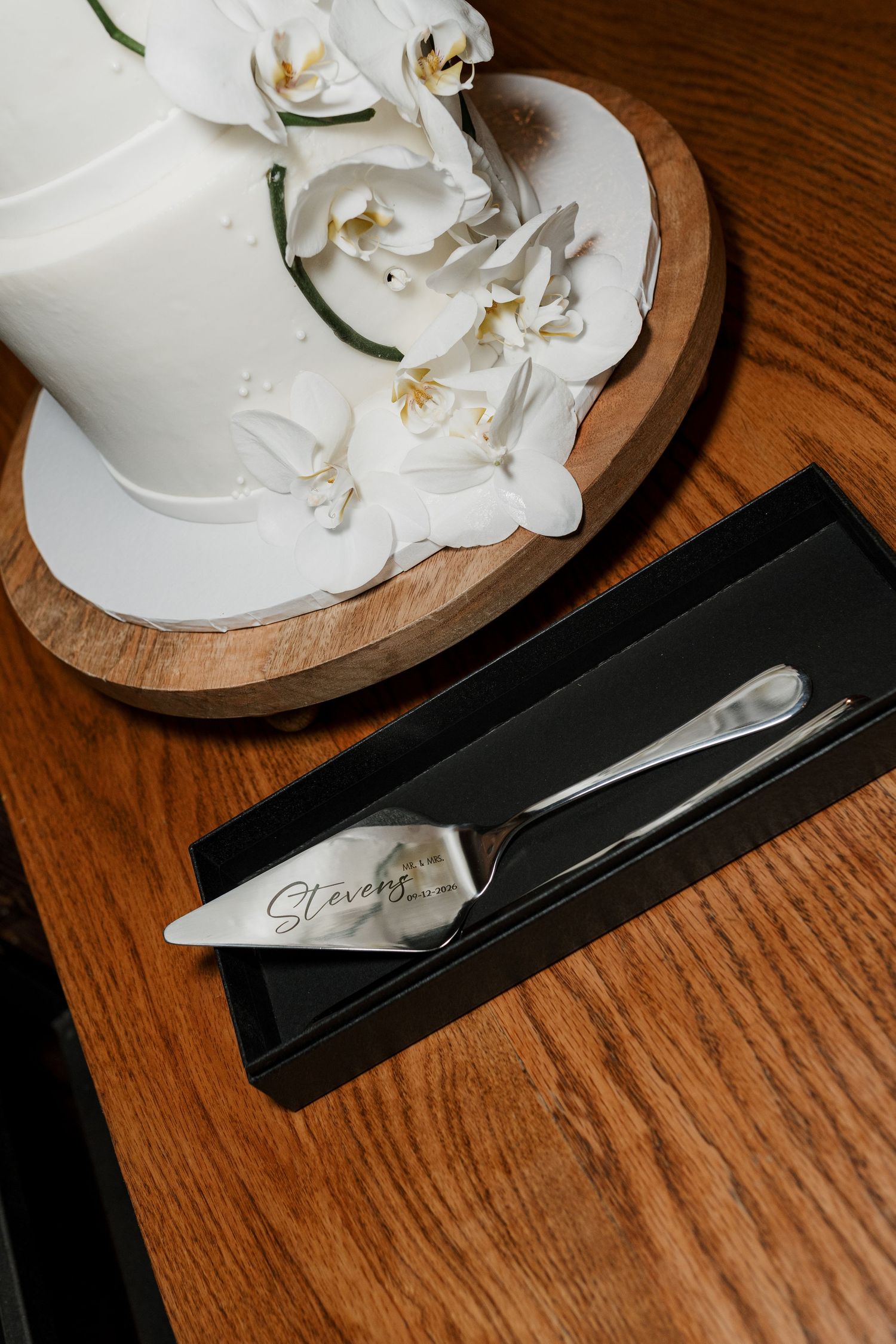 White wedding cake with orchids on wood stand beside personalized Stevens cake server set in black gift box.
