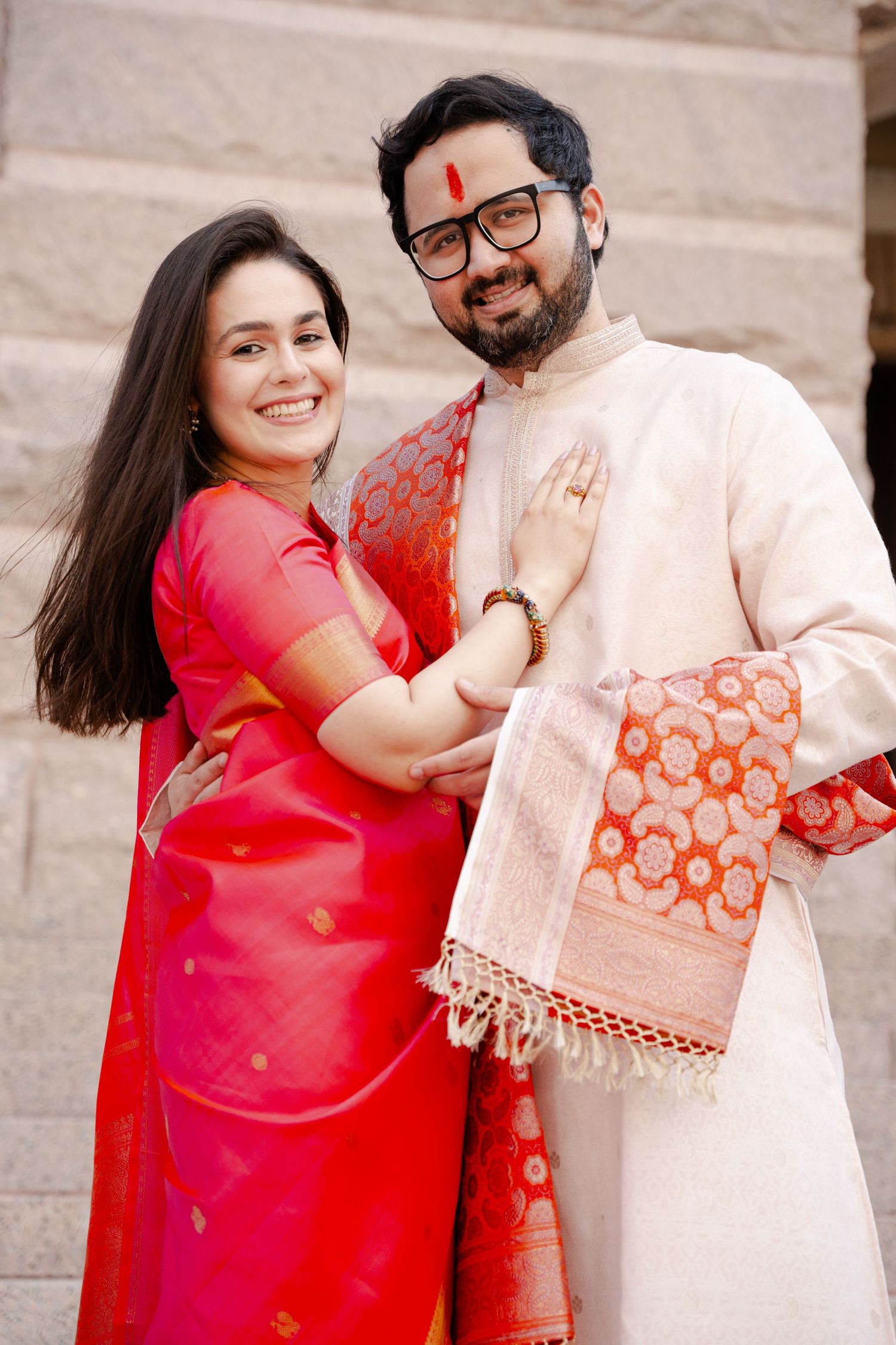 A couple in traditional Indian attire poses together, wearing red and cream colored outfits.