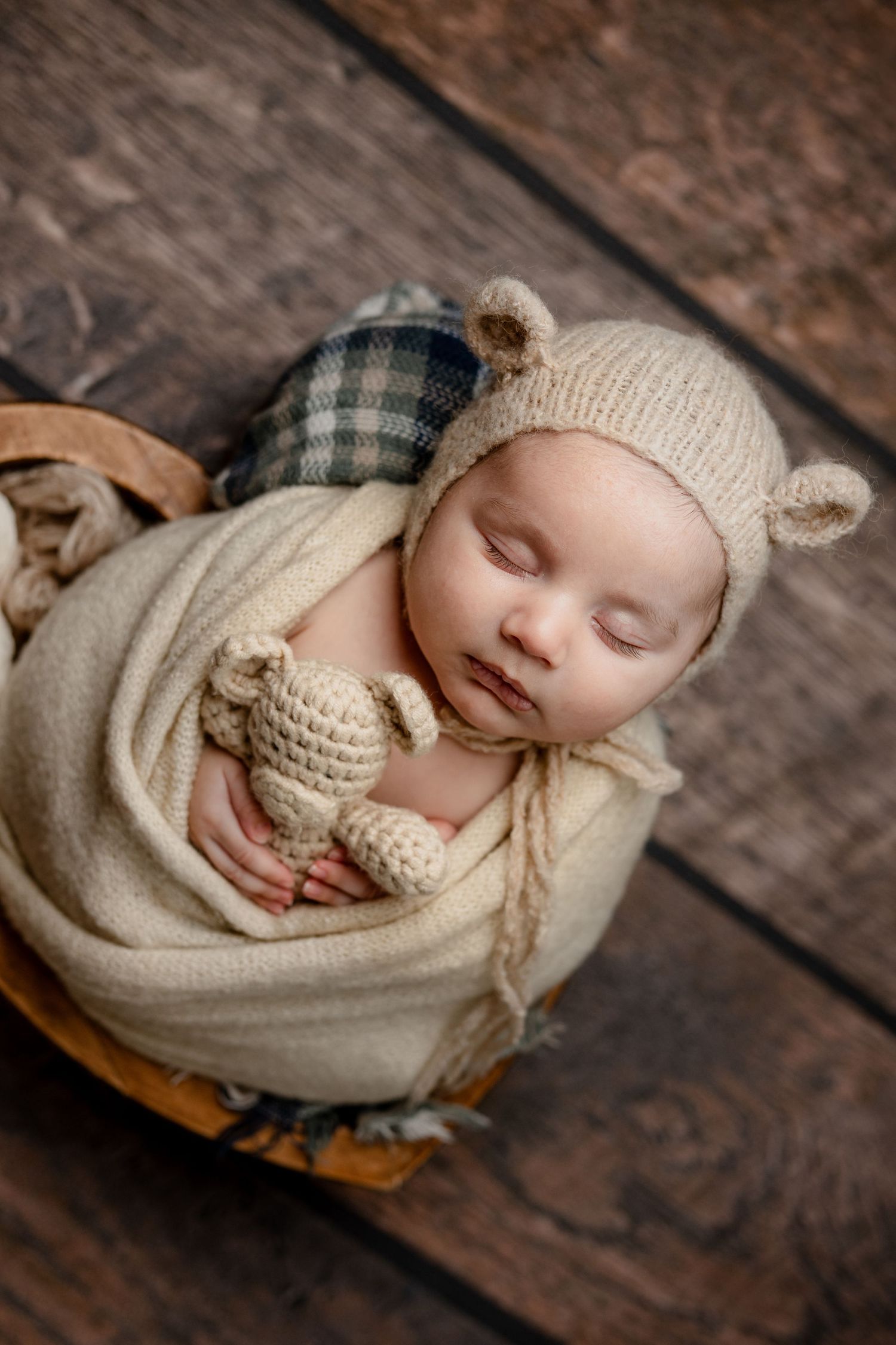 Sleeping newborn wrapped in cream knit blanket wearing bear ear bonnet and holding small teddy bear on wooden floor.