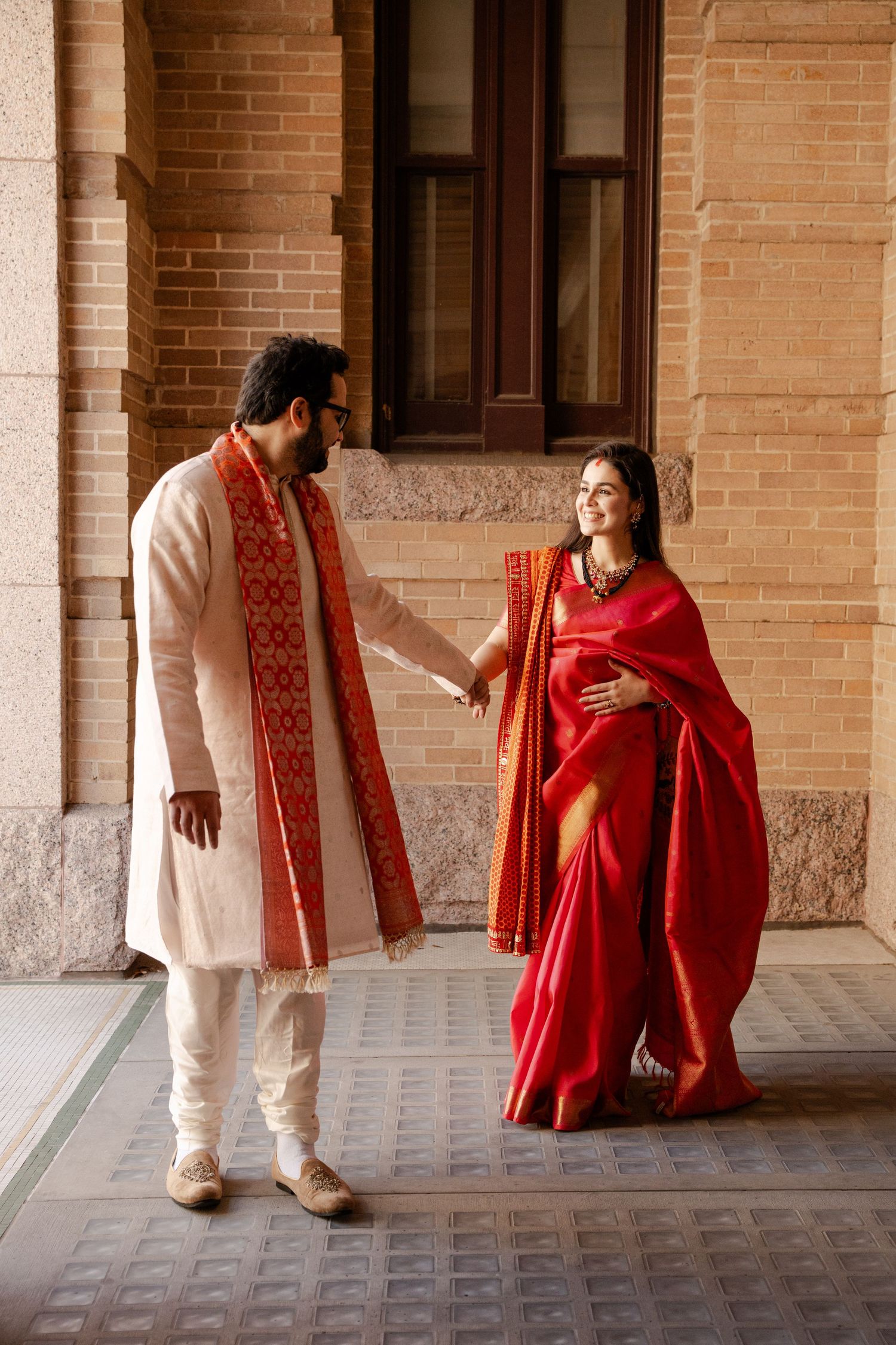 A couple in traditional Indian wedding attire sharing a moment in an ornate brick corridor.