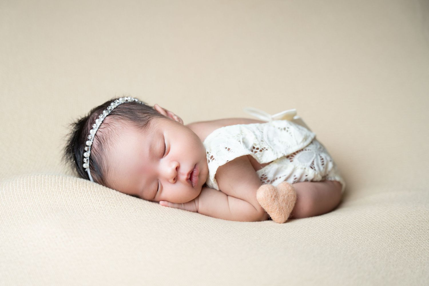 Sleeping newborn wearing white lace dress and sparkly headband rests peacefully on beige blanket.