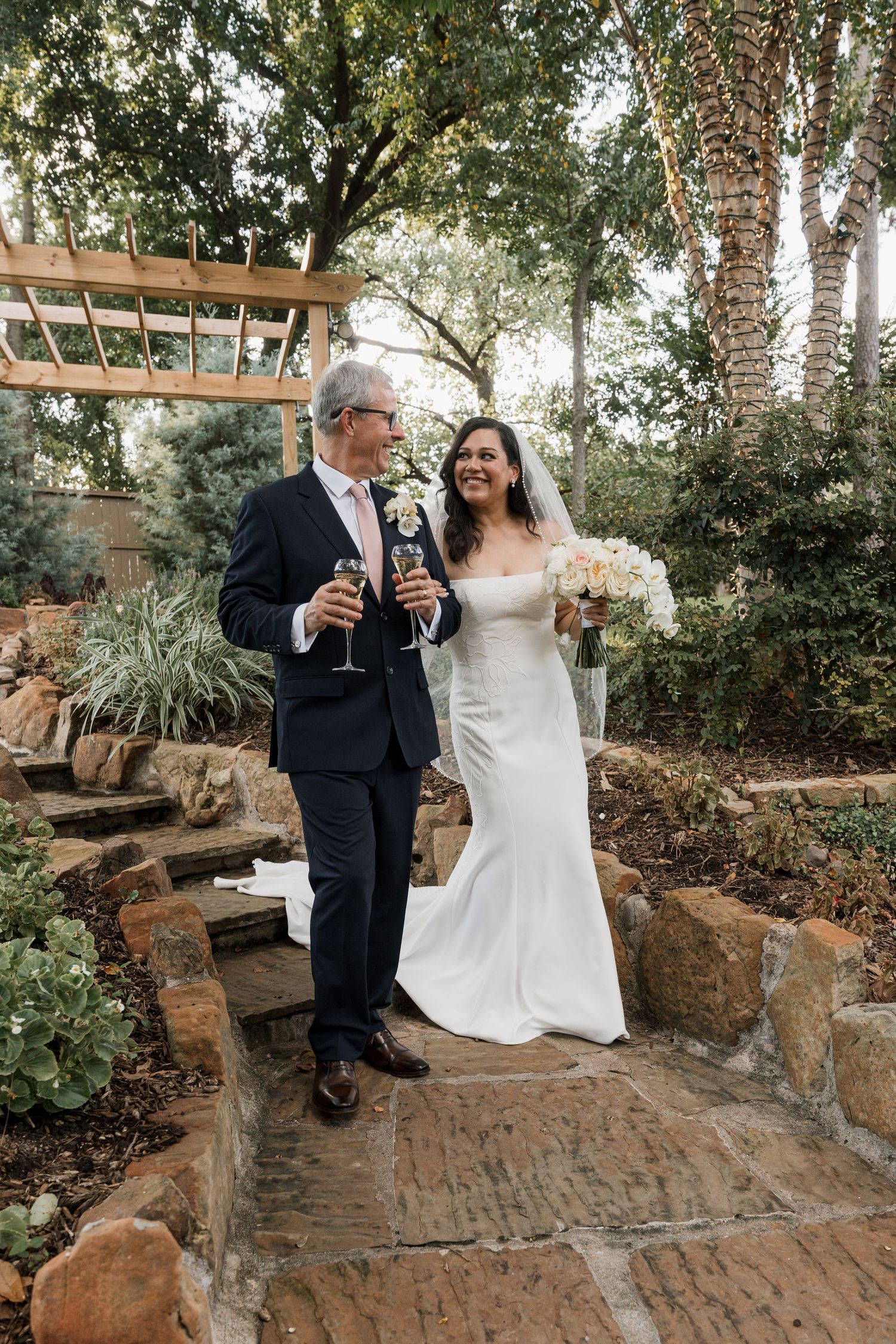 Bride and groom toasting with champagne glasses in a lush garden setting, bride holding white bouquet.