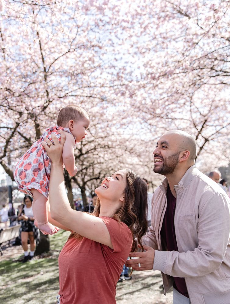 A husband, wife, and their daughter laughing in front of the Cherry Blossoms in Portland, OR.