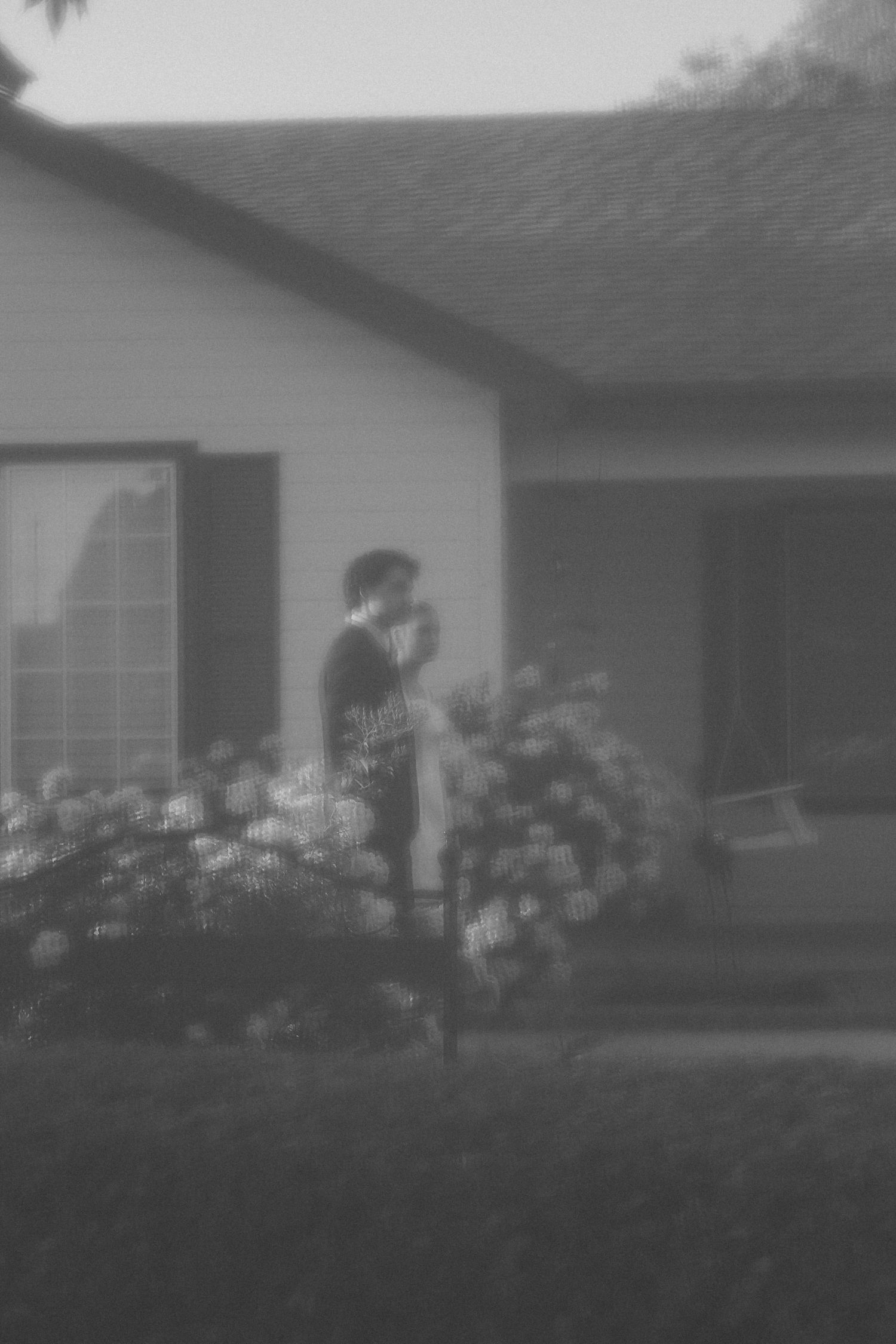 Blurry black and white photo of a person standing in front of a house surrounded by blooming hydrangea bushes.