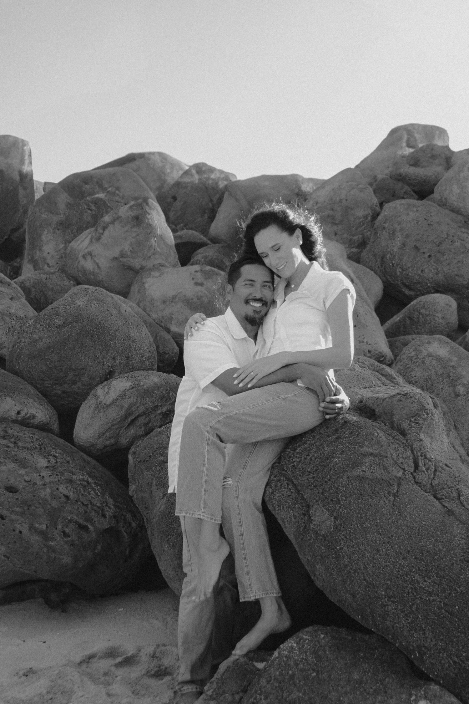 A loving couple embraces while sitting on large coastal rocks in a black and white beach photo.