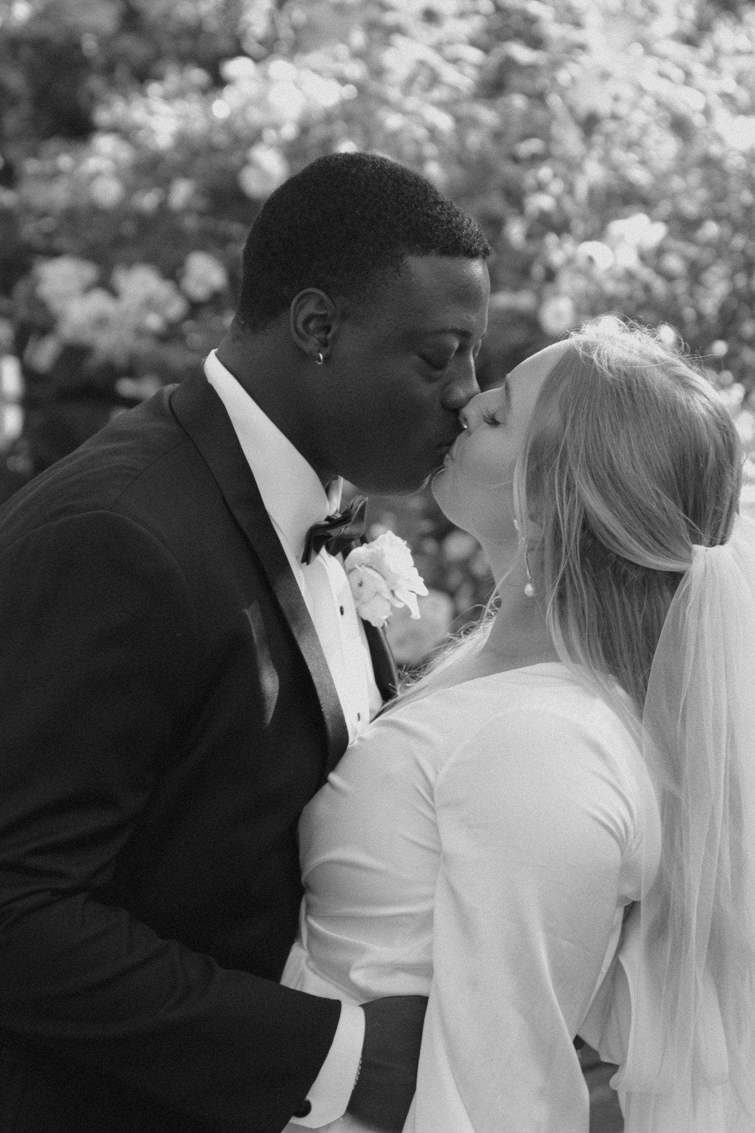 Black and white wedding photograph showing a couple sharing a romantic kiss in formal attire with a veil and flowers.