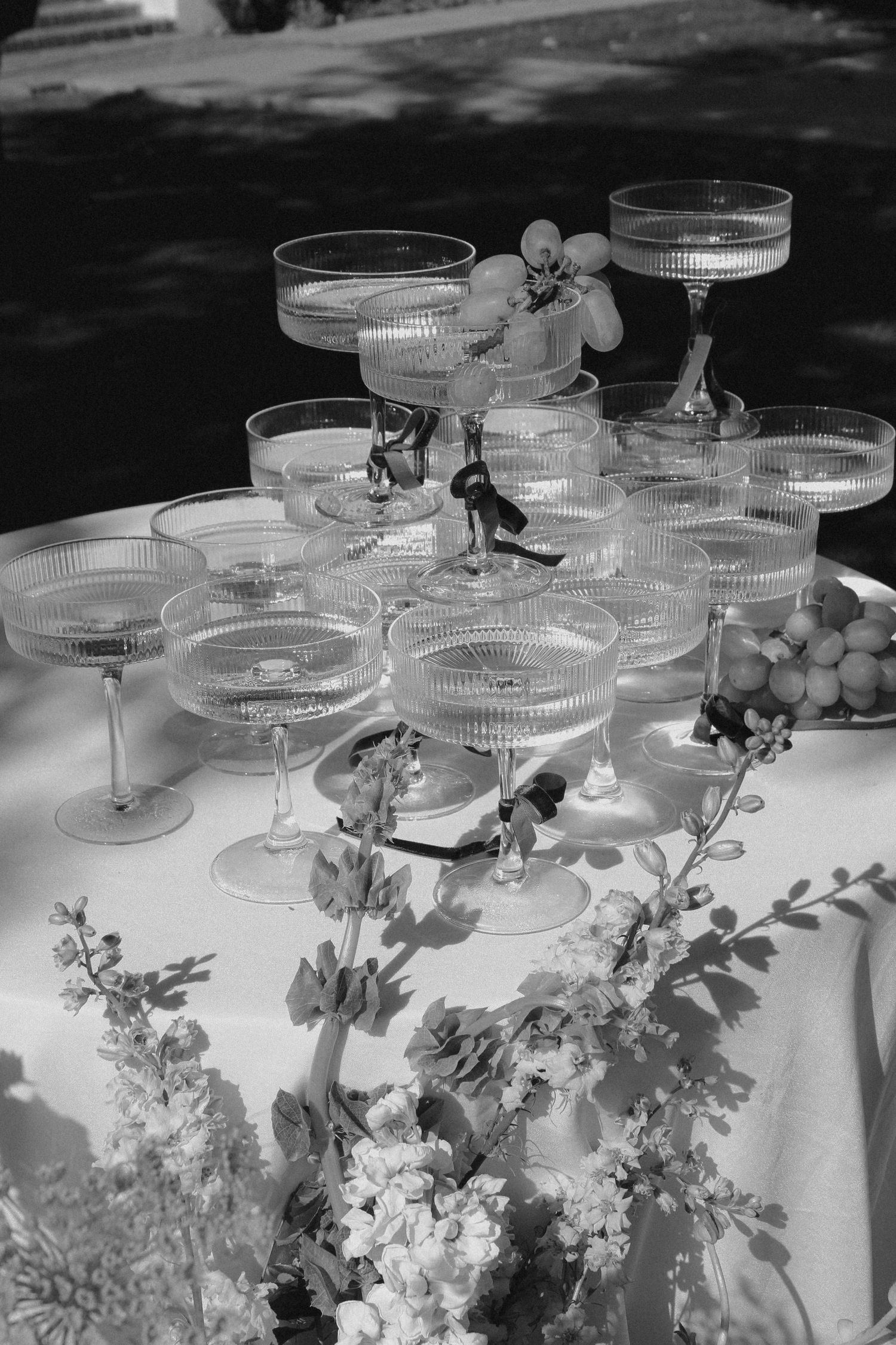 Black and white photo of vintage champagne coupes stacked in a pyramid with delicate floral arrangements on a tablecloth.