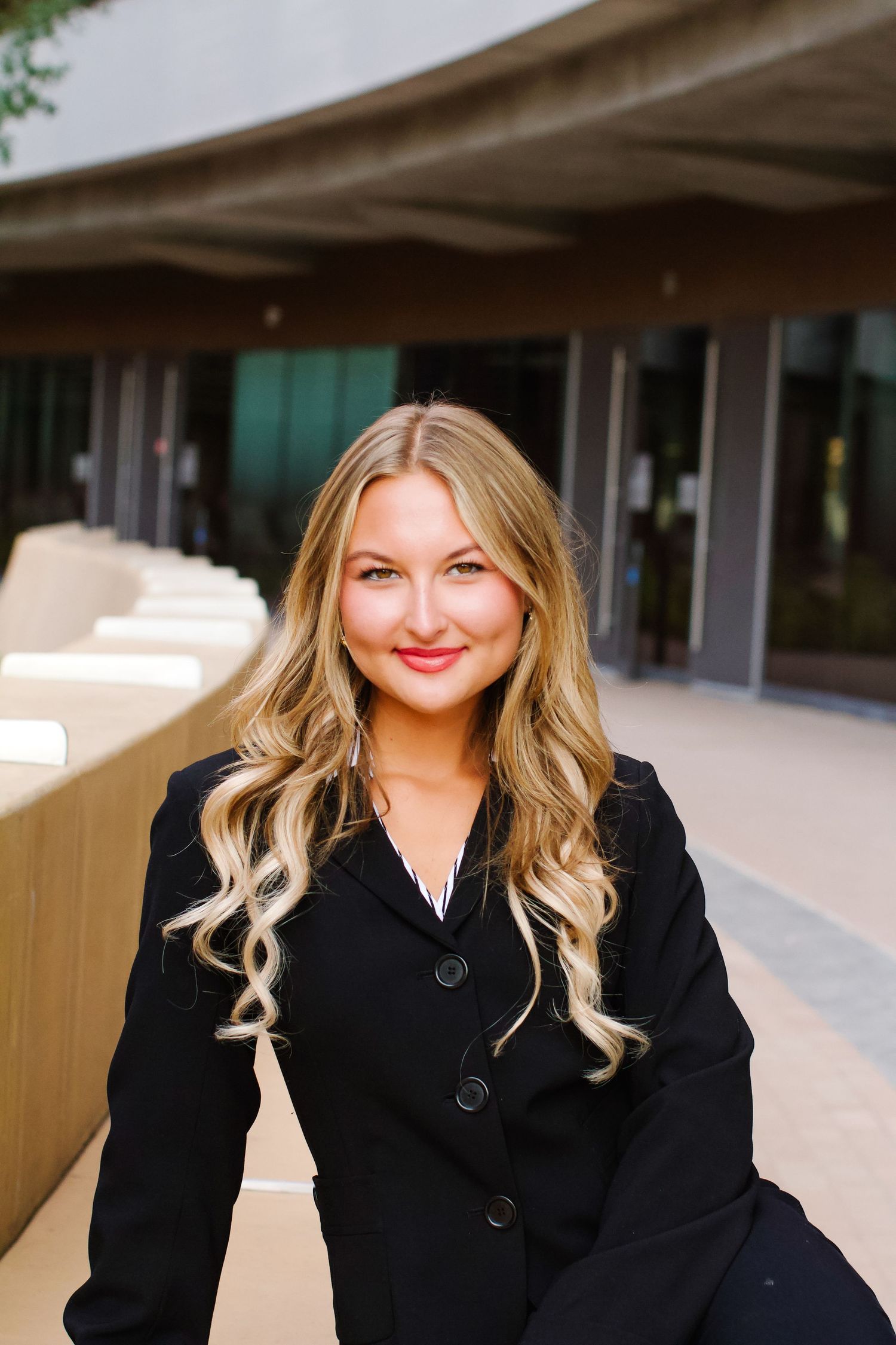 Professional portrait of a person in a black blazer smiling against a modern building exterior.