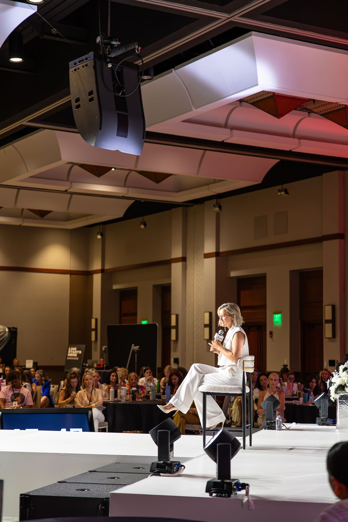 Woman in white outfit sits on stage speaking to a large conference audience in a modern ballroom.