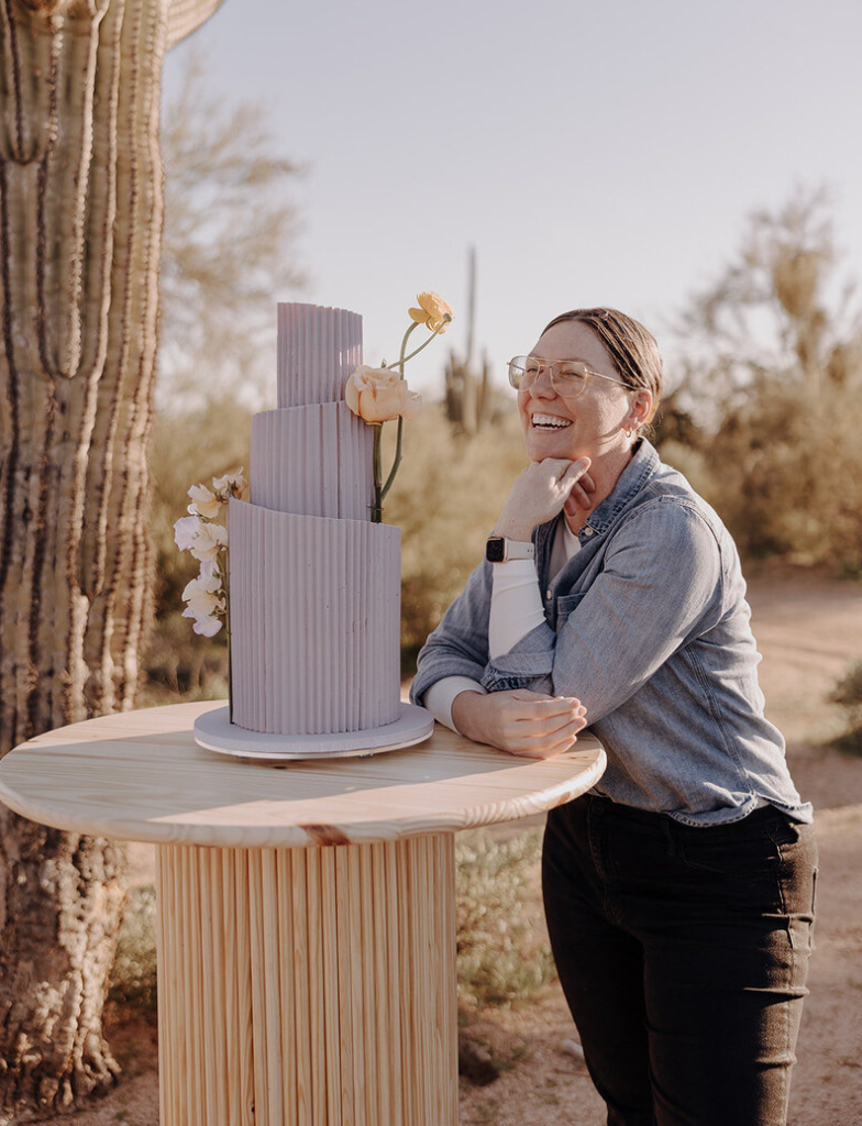 A chef making decorative cuts and patterns into a circular cake on a wooden pedestal stand in a rustic setting.