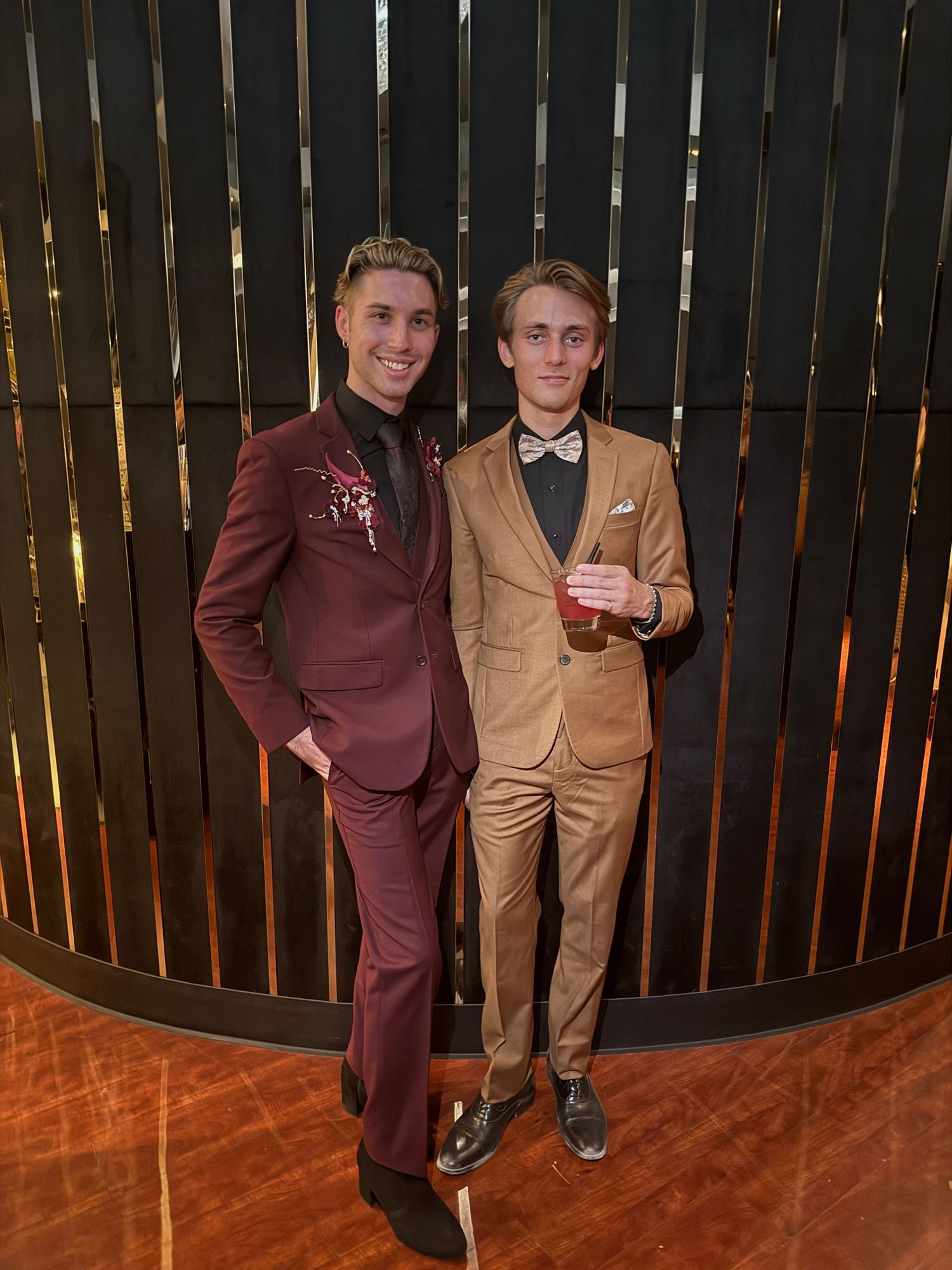 Two young men in formal suits, one maroon and one tan, posing together at an elegant event venue.