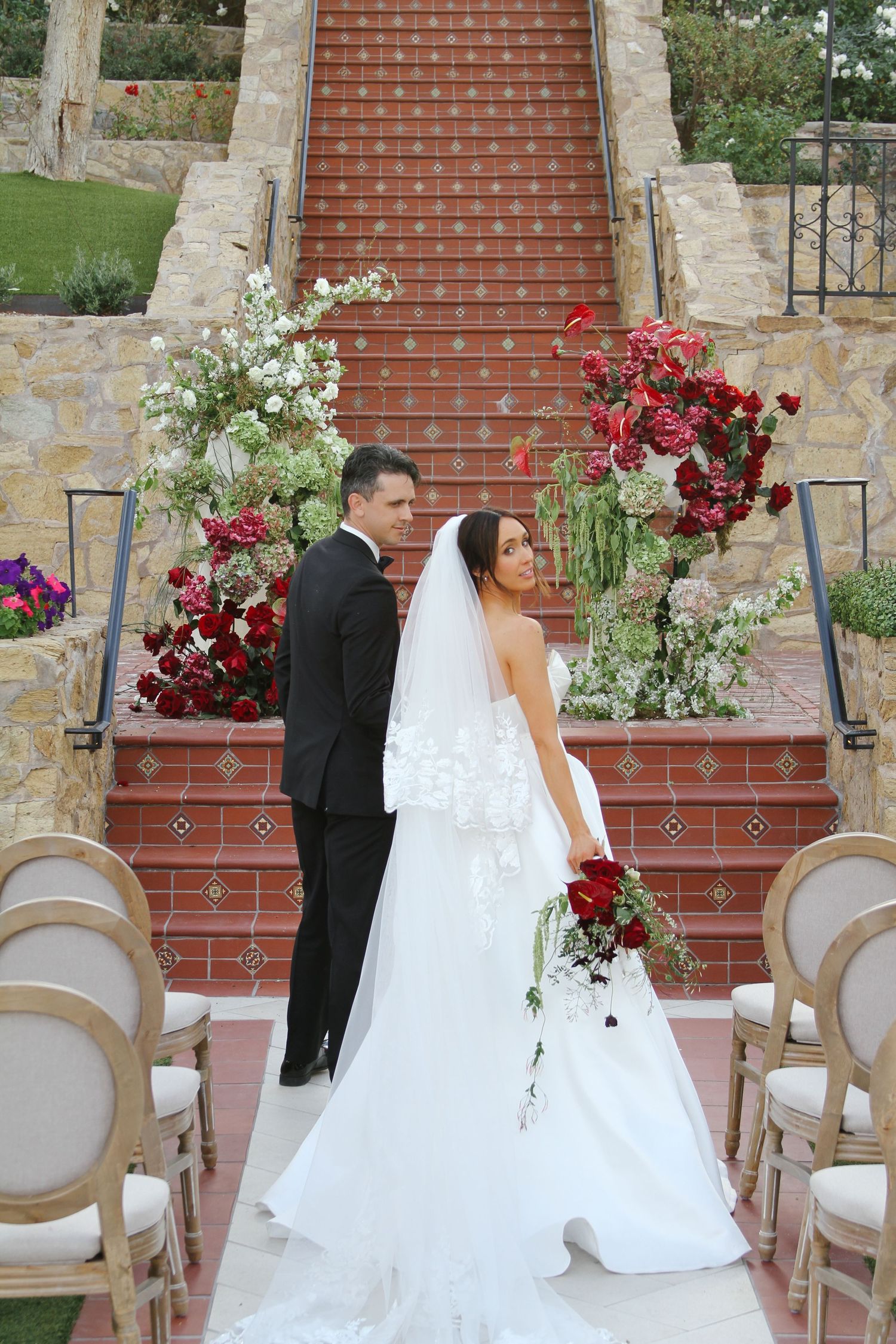 Bride and groom pose together at outdoor wedding ceremony on red-tiled stairs adorned with lush red and white floral arrangements.
