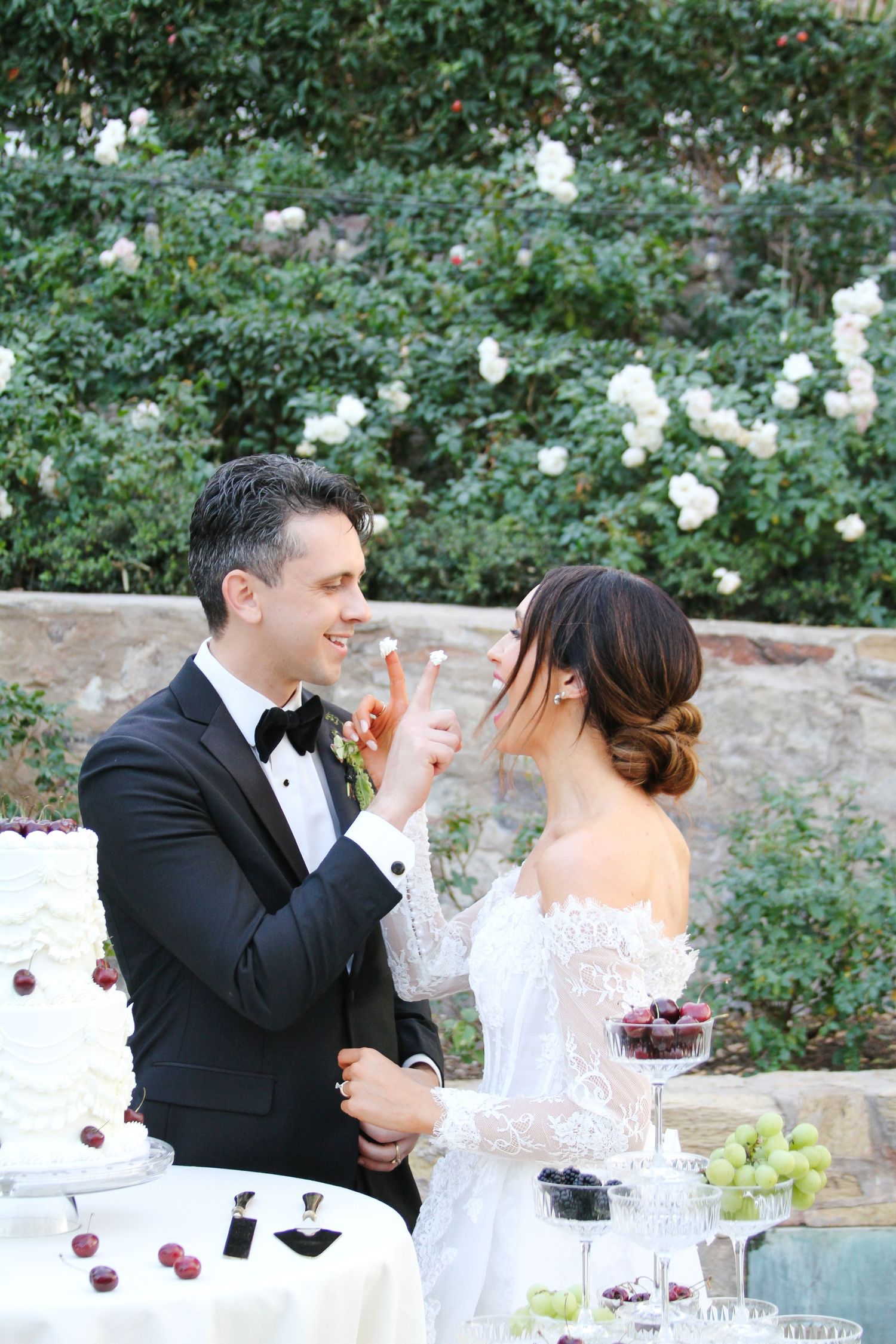 Groom in tuxedo feeds bride a cherry at elegant outdoor wedding reception with white cake and floral backdrop.