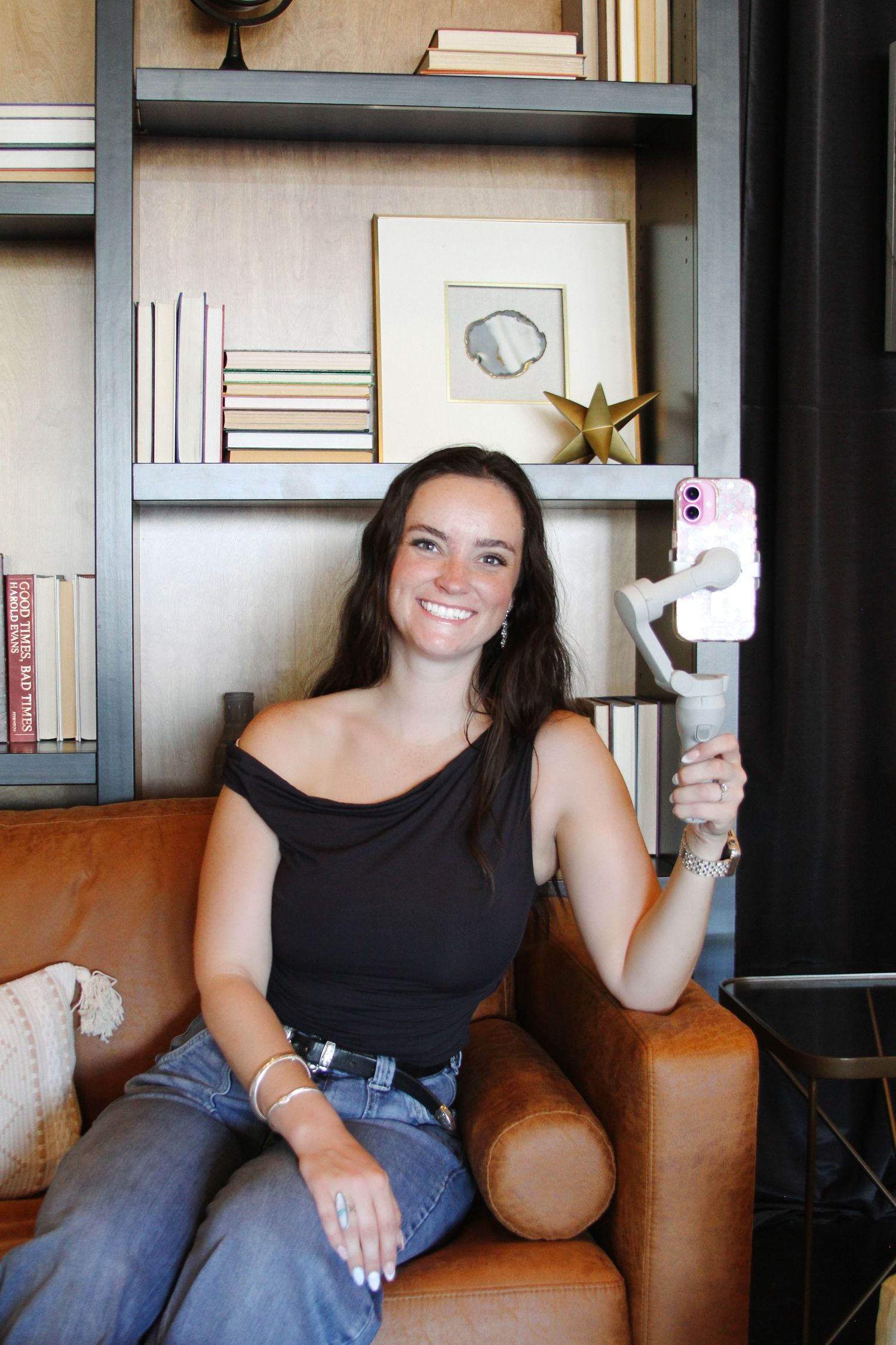 A woman sitting on a leather chair holds up a phone while smiling in front of a bookshelf.