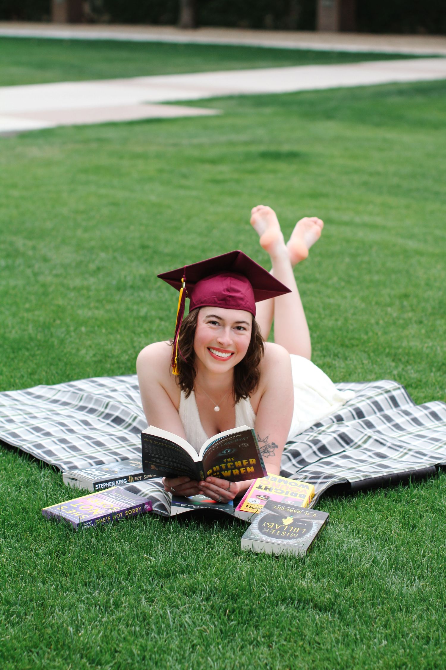 Graduate in red cap and yellow stole lying on blanket with books on grass.