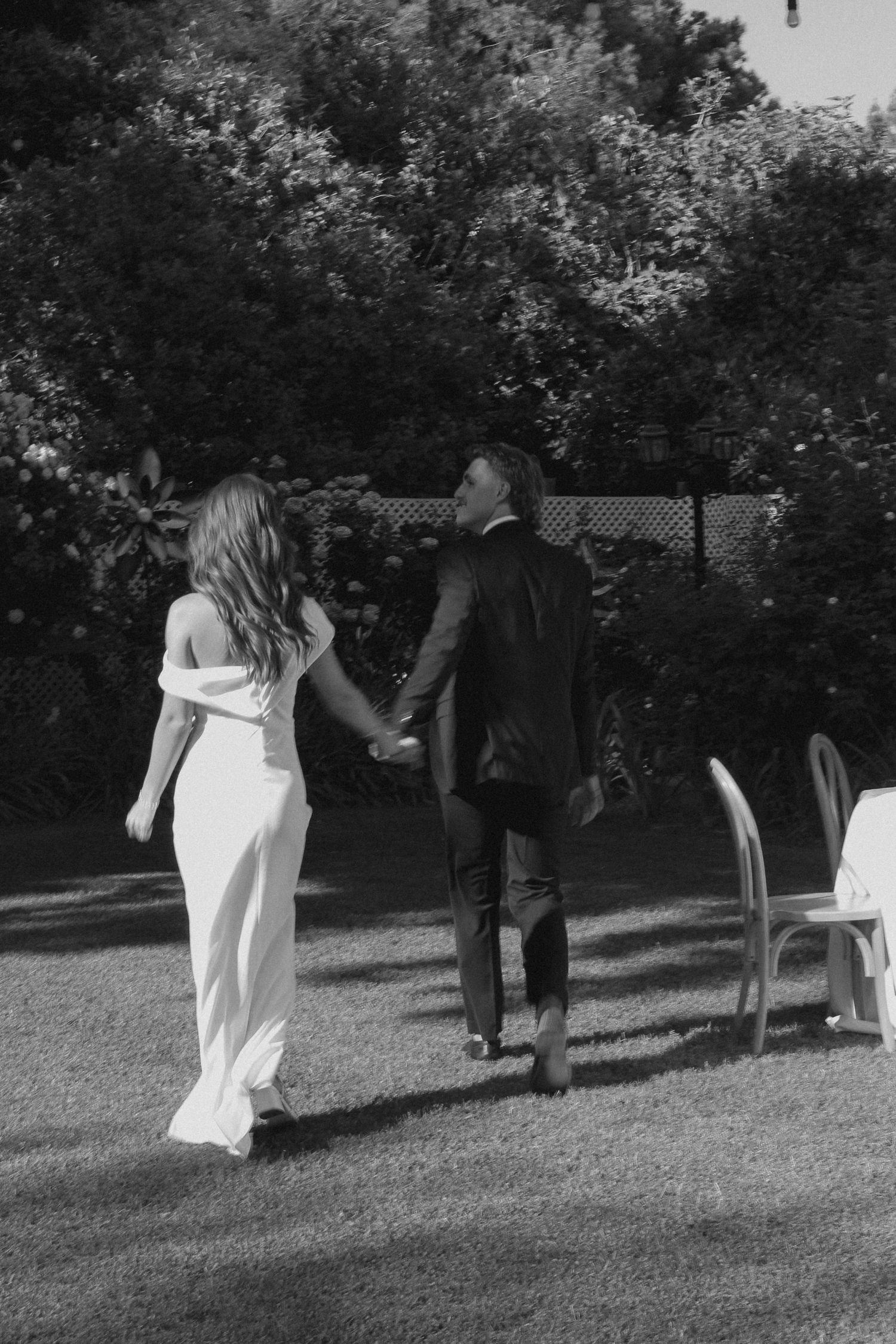 Black and white photo of a couple holding hands while walking on grass in a garden setting.