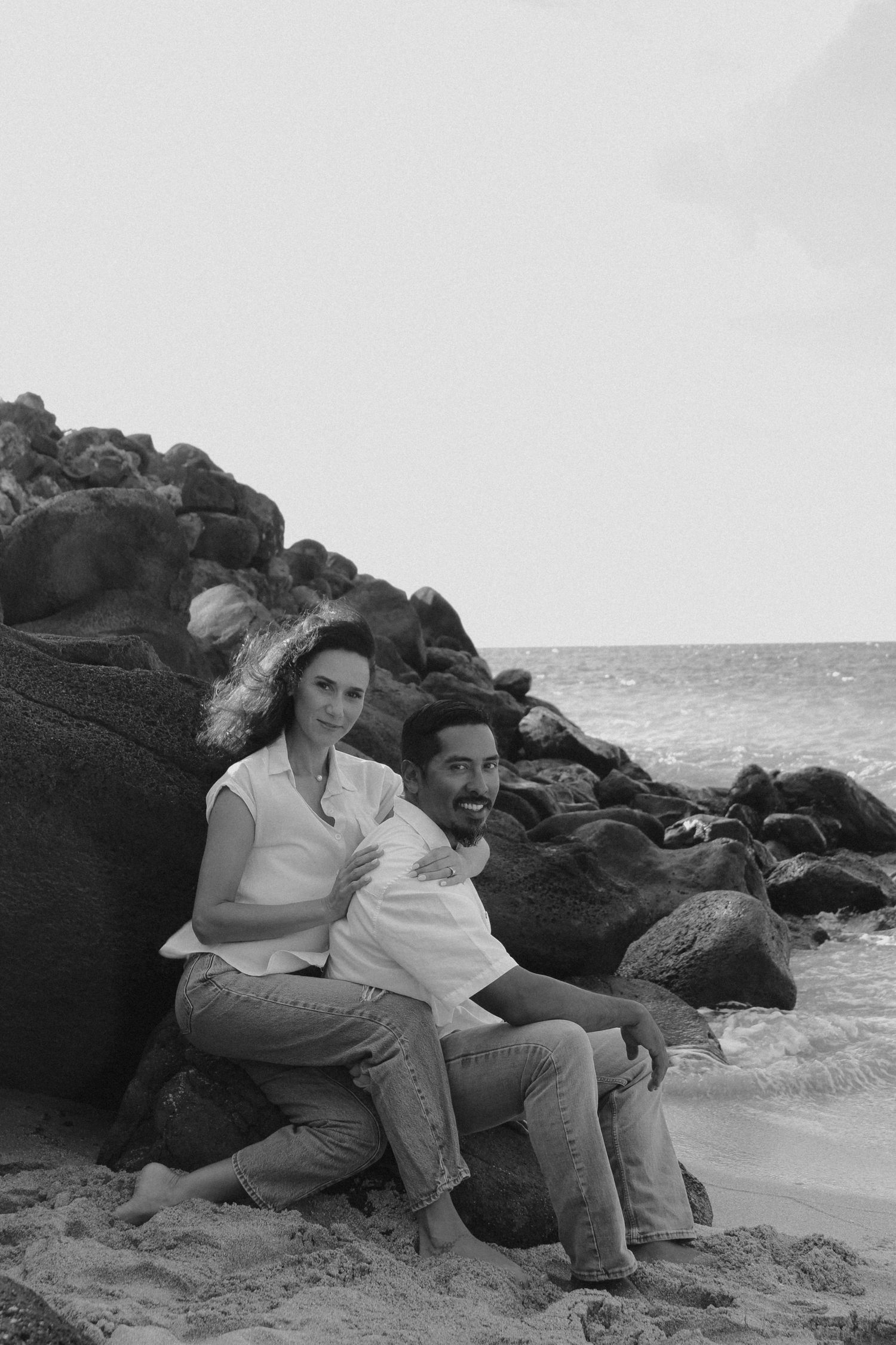 A couple relaxes together on rocky beach shore in black and white photograph.