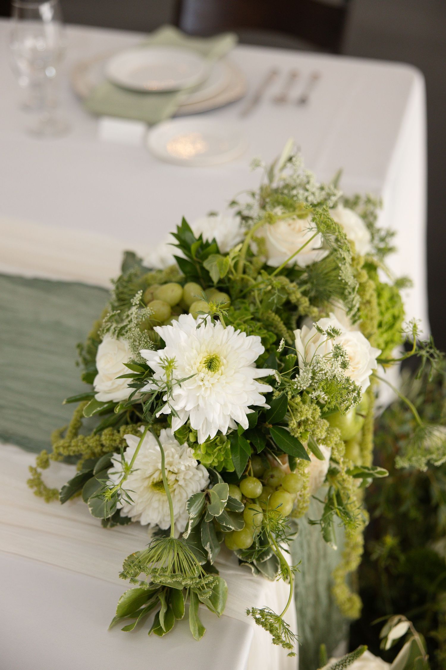 Lush white floral wedding centerpiece with green grapes and foliage cascading over a white tablecloth.