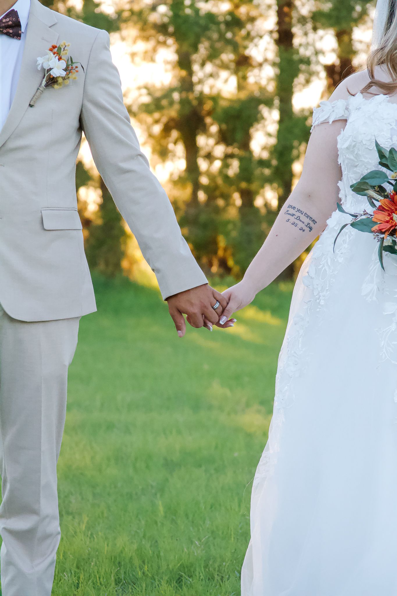 A couple in wedding attire holds hands during sunset on a grassy lawn, with the groom in light suit and bride in white dress.